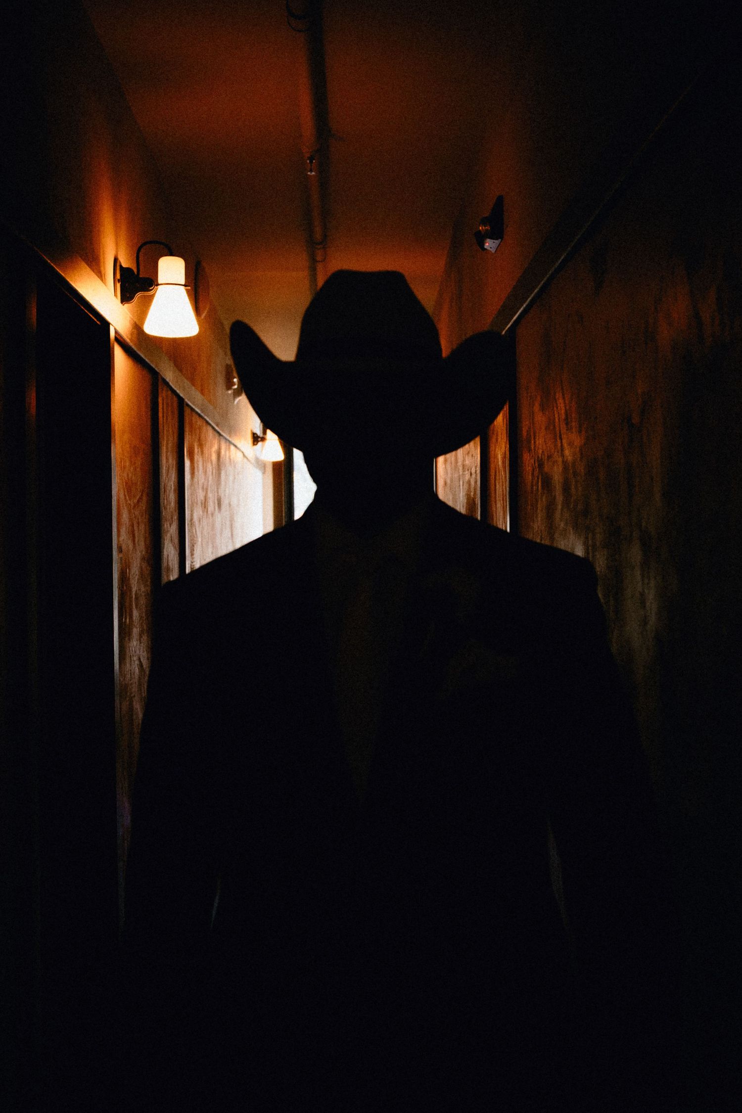 Silhouette of a person wearing a cowboy hat standing in a dimly lit corridor with warm glowing wall lights.