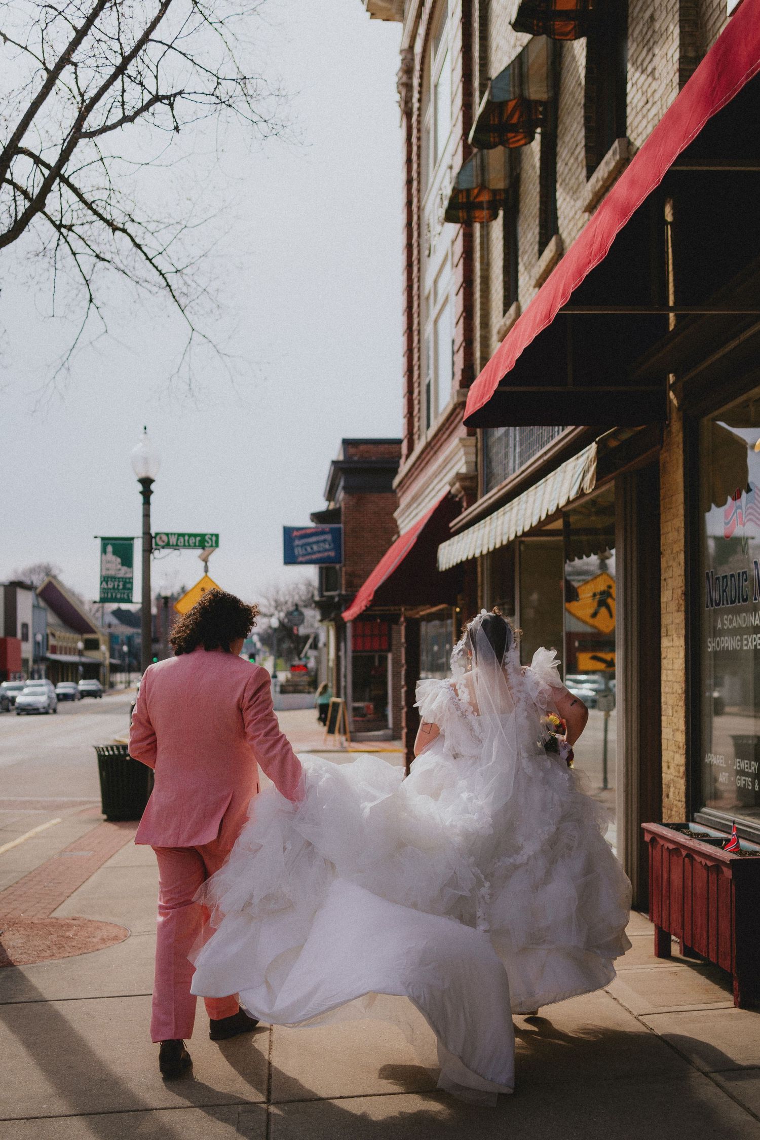 Bride in flowing white gown and groom in pink suit walking along a downtown sidewalk on their wedding day.