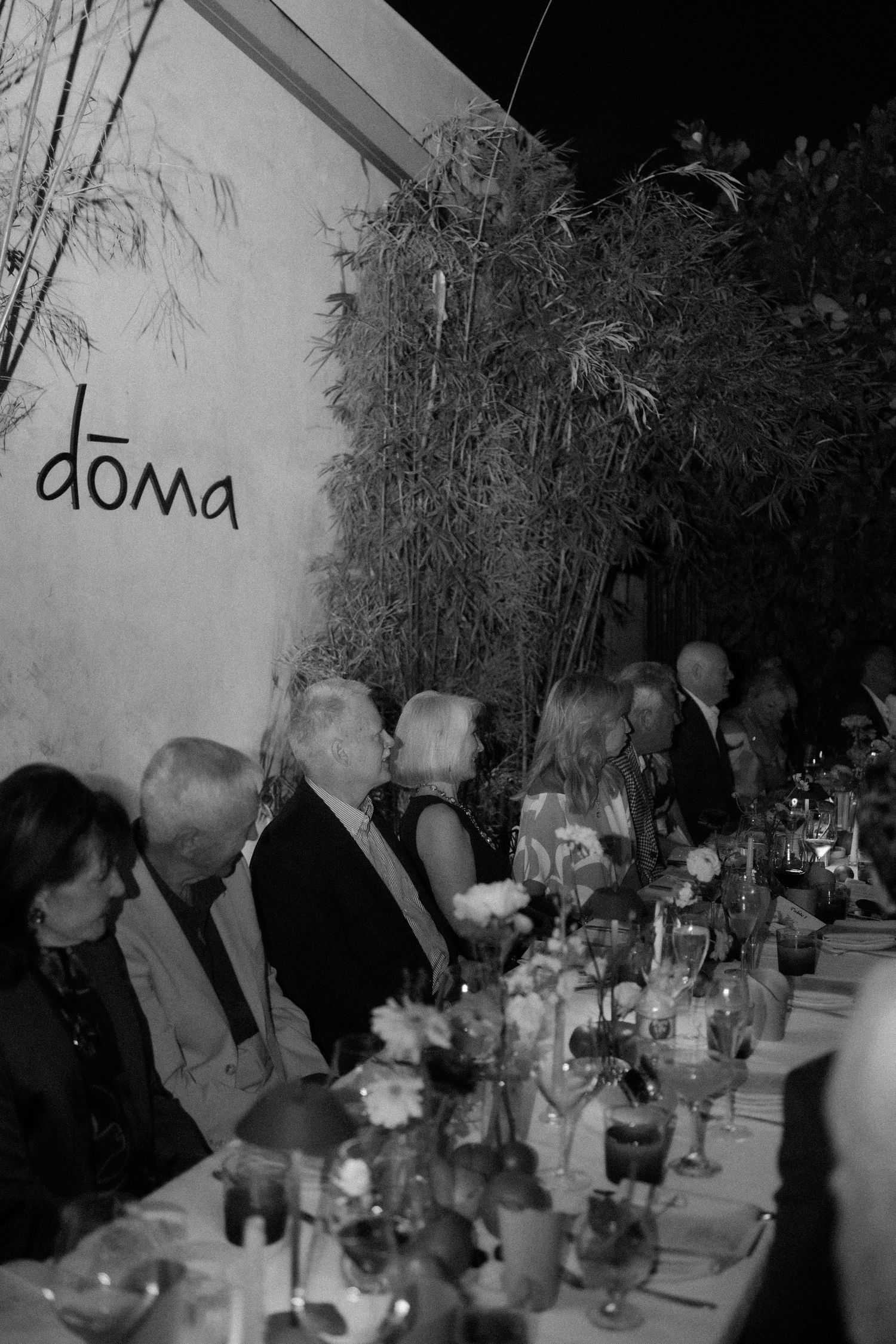 Black and white photo of elegant dinner party guests seated at a long table at Doma restaurant at night.