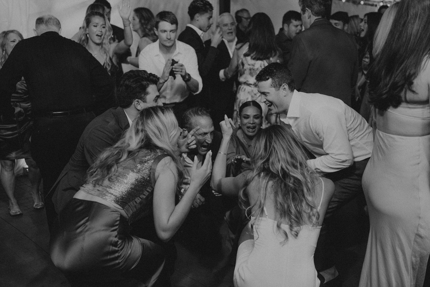 Black and white photo of wedding guests dancing and crouching together on crowded dance floor.
