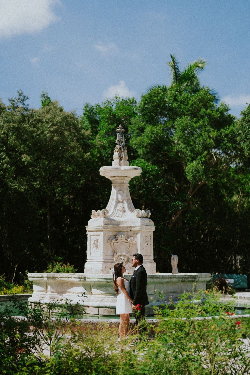 A couple standing near an ornate white stone fountain in a lush garden setting.