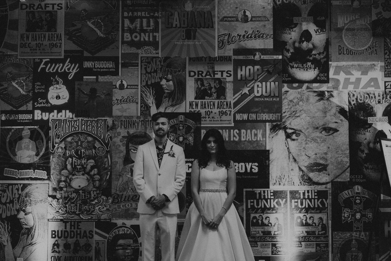Bride and groom pose before a wall of vintage concert posters in a stylish black and white wedding photo.