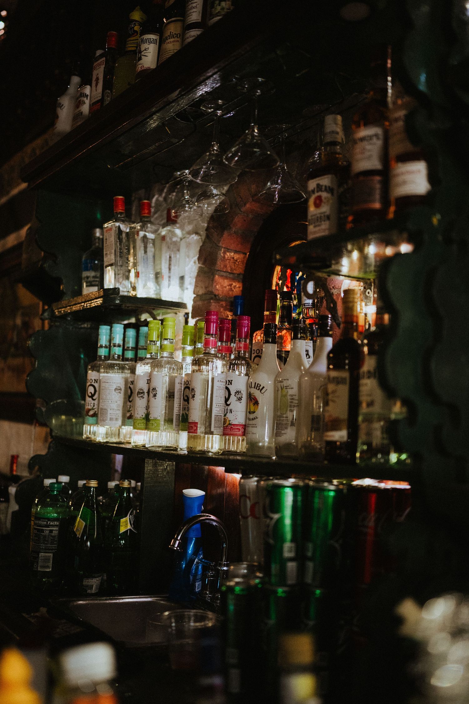 Dimly lit bar shelves stocked with various liquor bottles including Bacardi rum and vodka in a moody nightclub setting.
