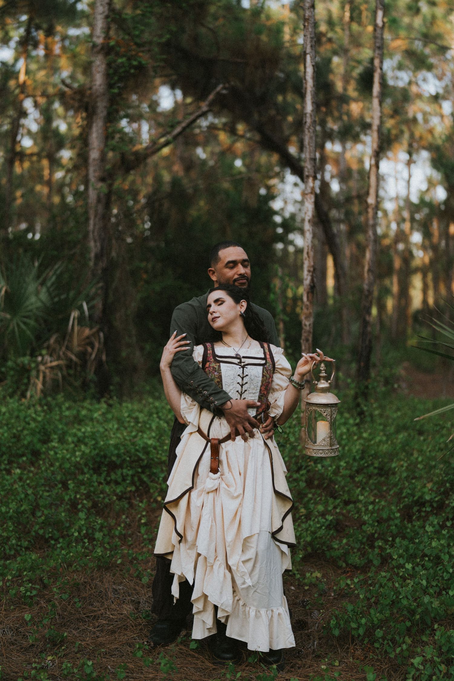 Couple in medieval fantasy costumes embracing in forest, woman in cream ruffled gown holding lantern, man in dark outfit behind her.