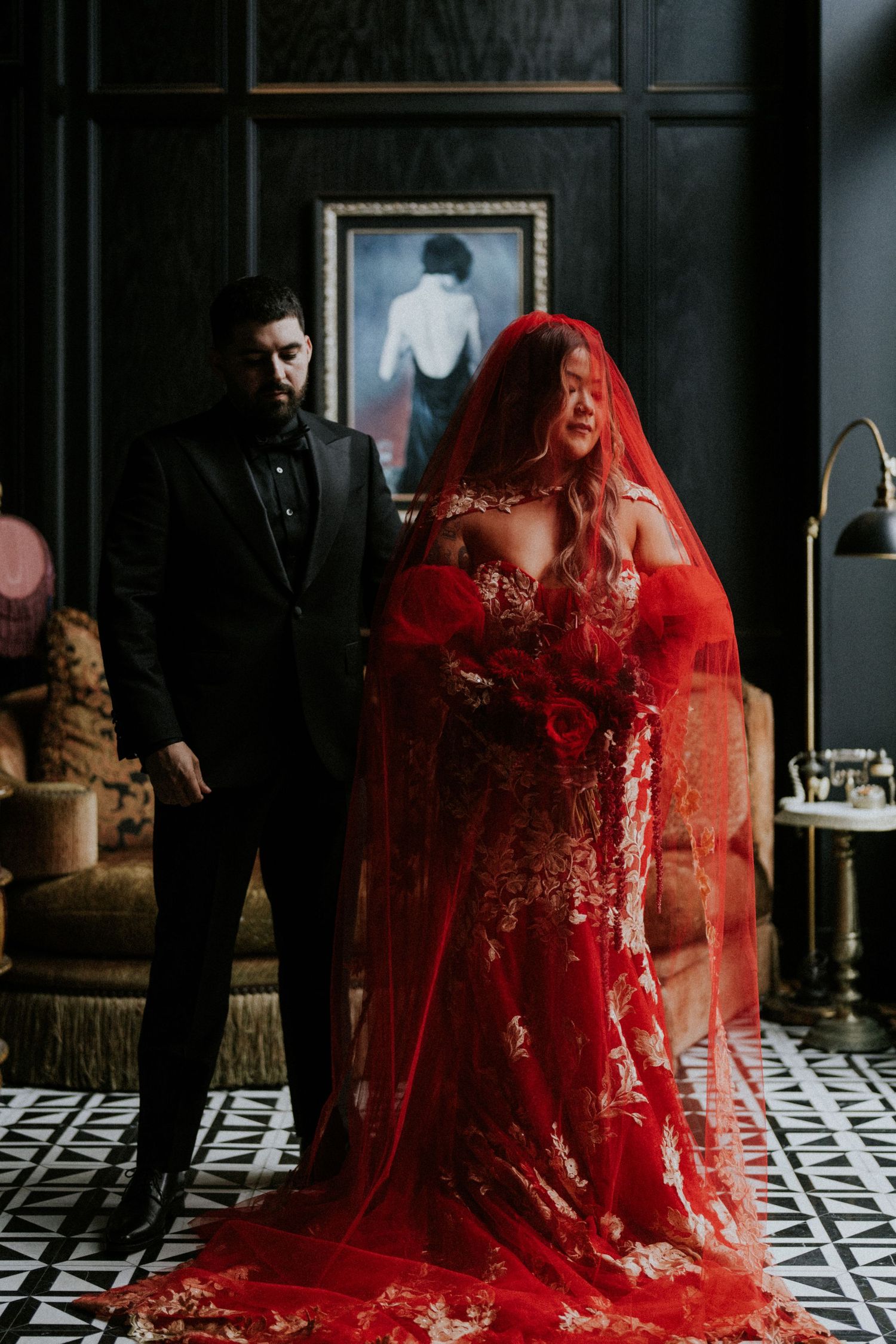 Couple in dark moody room, bride in red lace gown with veil, groom in black suit, gothic wedding style.