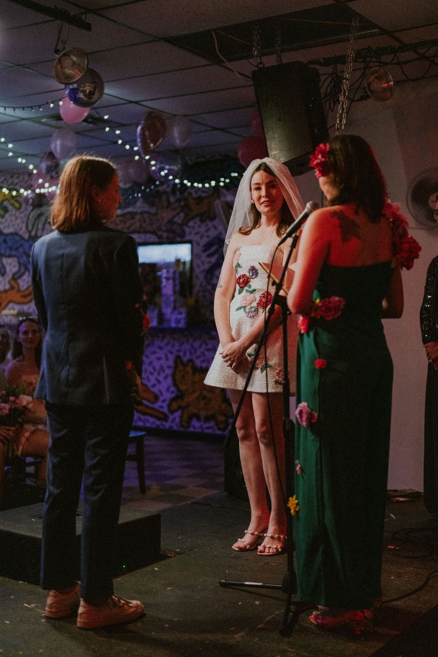 Bride in floral dress and veil stands at microphone during indoor wedding ceremony with guests.