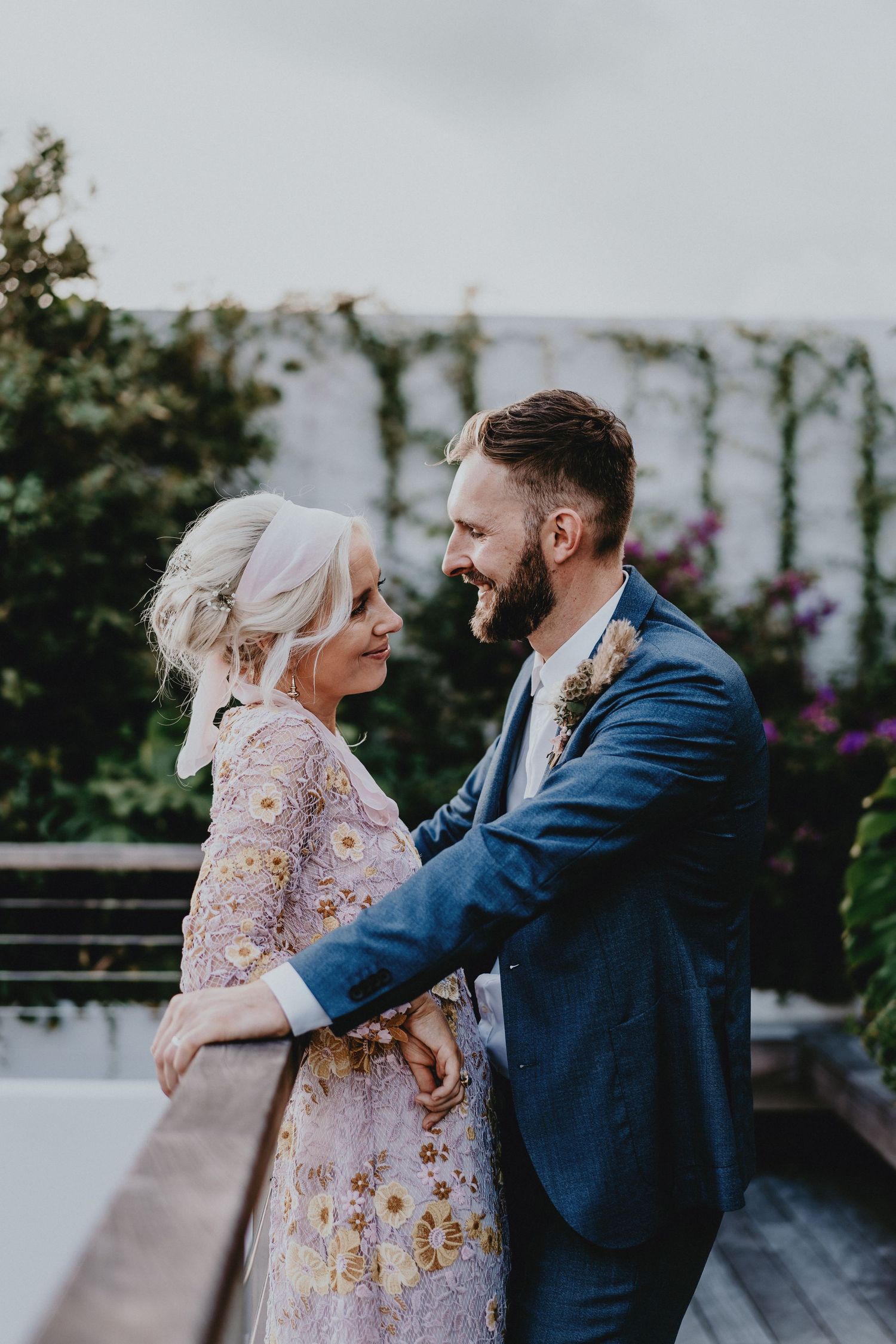 A couple in wedding attire share a romantic moment on a garden balcony, the bride in floral dress and groom in navy suit.