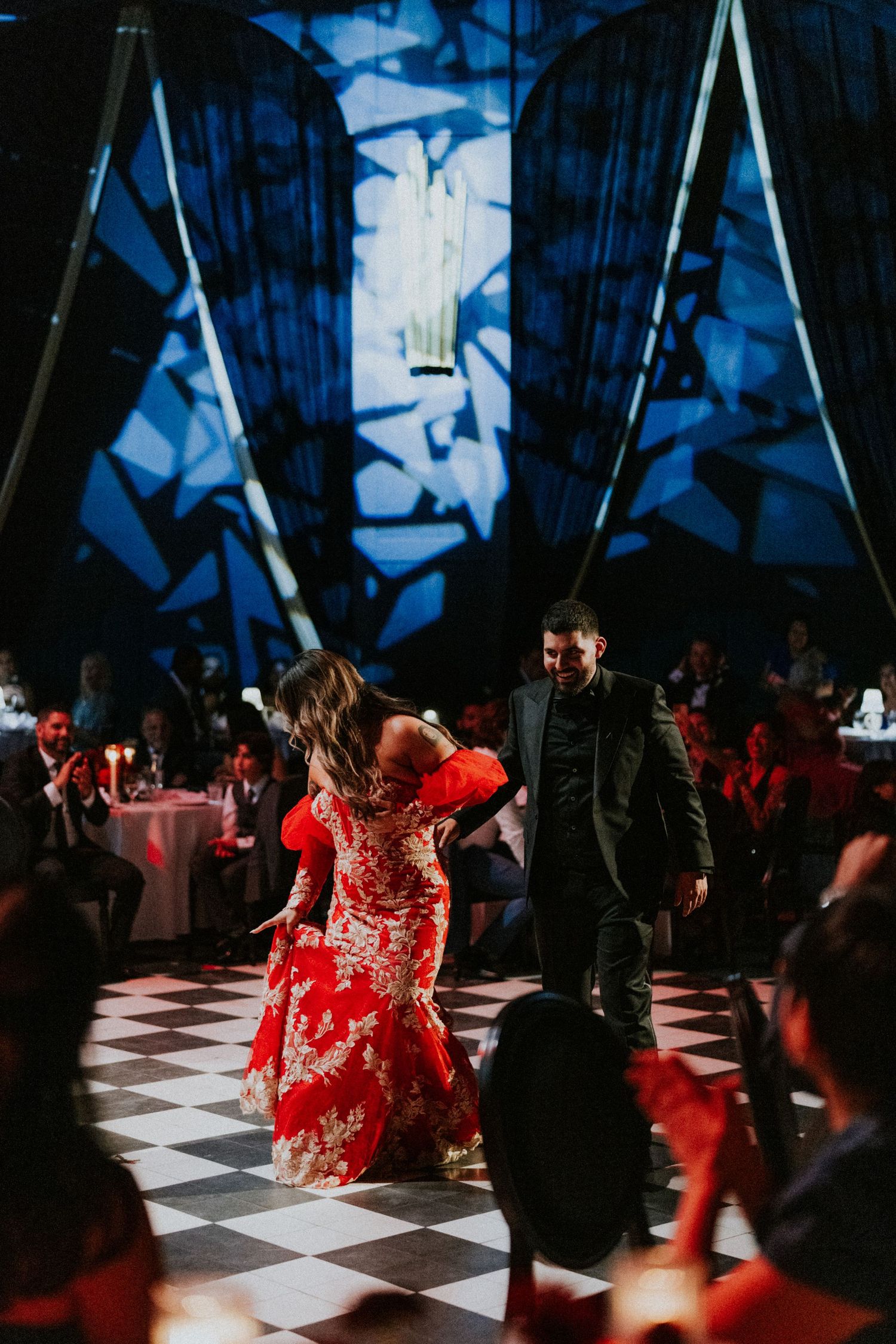 Couple dancing on checkered floor in elegant venue with blue geometric lighting projections and red floral gown.