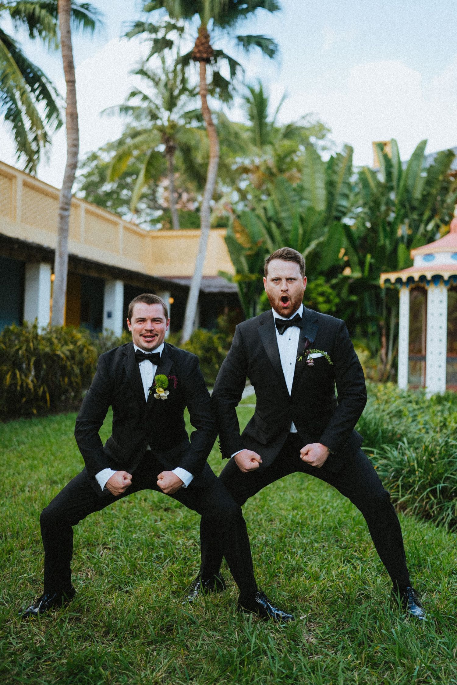 Two groomsmen in black tuxedos strike a fun pose on grass with tropical palm trees and a building in the background.