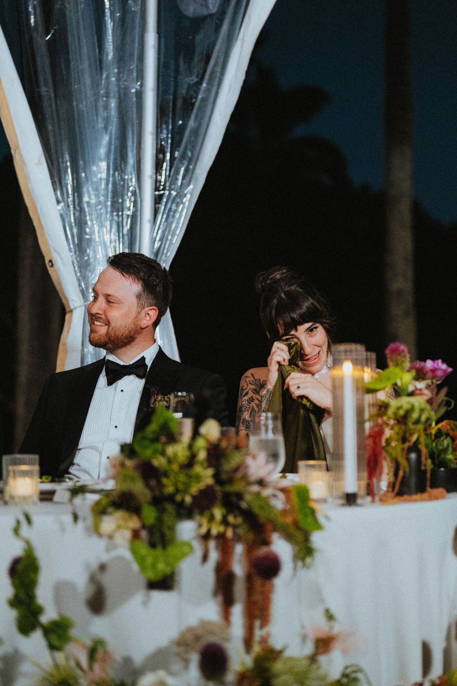 Bride and groom sit at their sweetheart table surrounded by lush floral arrangements and candles during wedding speeches.