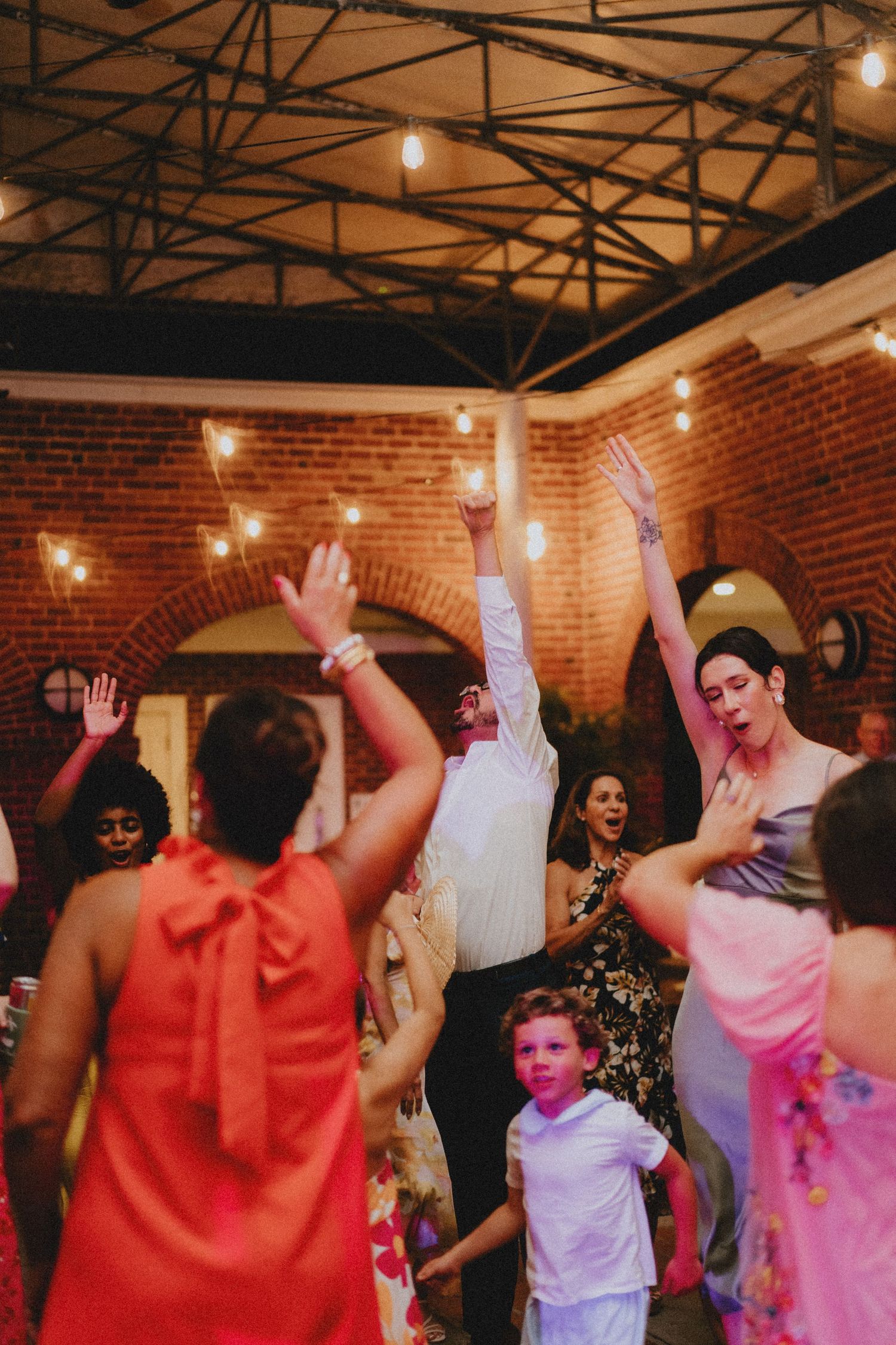 People dancing and celebrating at a wedding reception in a rustic brick venue with string lights overhead.