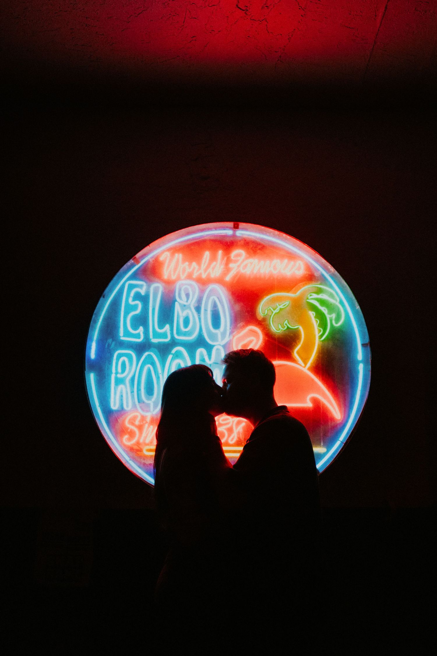 Silhouetted figures stand against a backdrop of colorful neon signs glowing in vibrant red, blue and green hues.