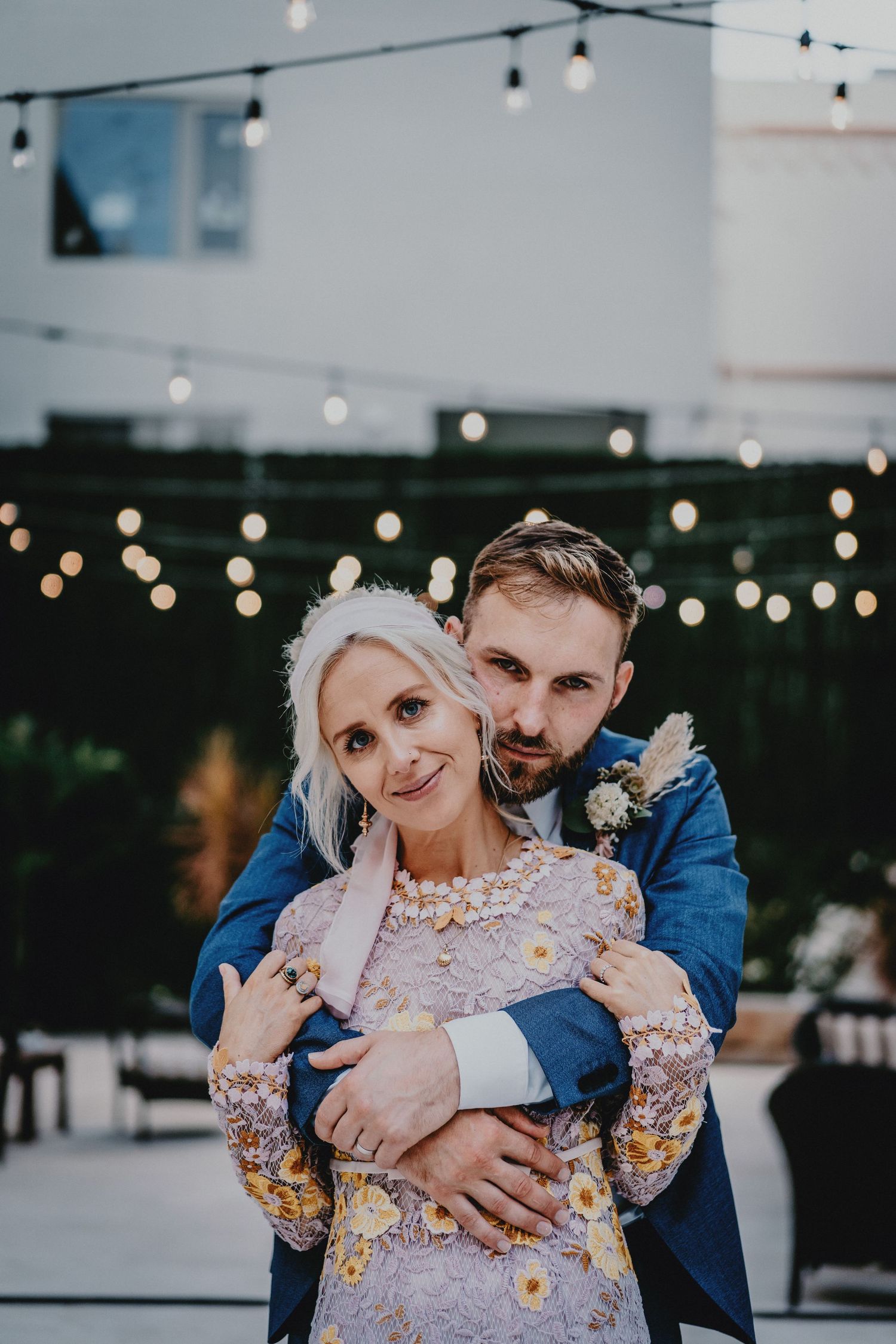 Couple embracing at outdoor wedding reception with string lights, groom in blue suit hugging bride in floral lace dress.