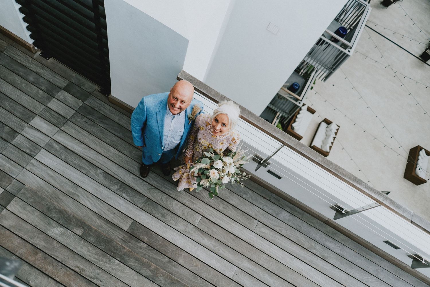 Aerial view of a wedding couple, groom in blue suit and bride holding a floral bouquet, standing on wooden deck.