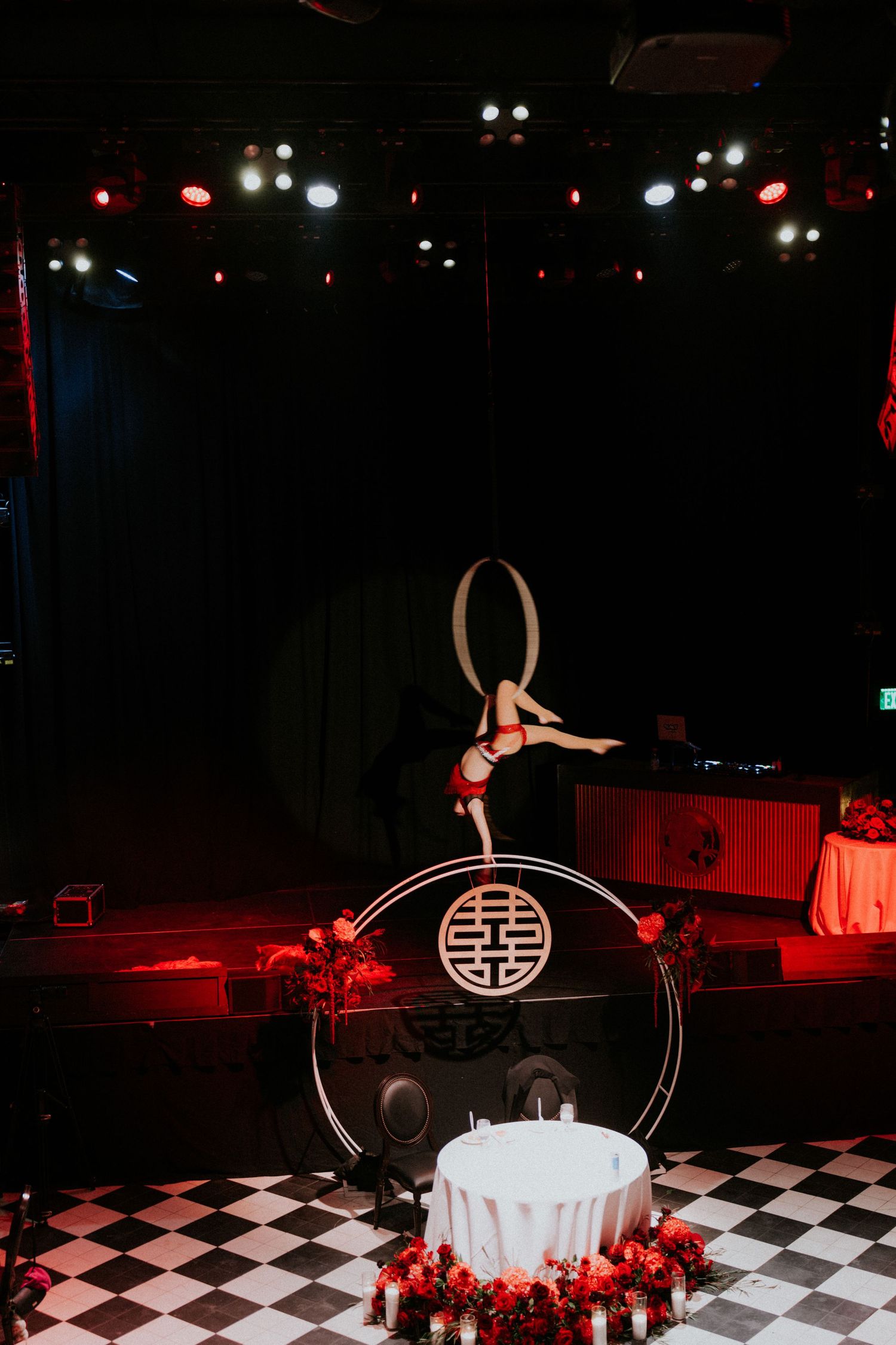 Aerial acrobat performs in a hoop above a Chinese symbol display on a checkered floor with red lighting.