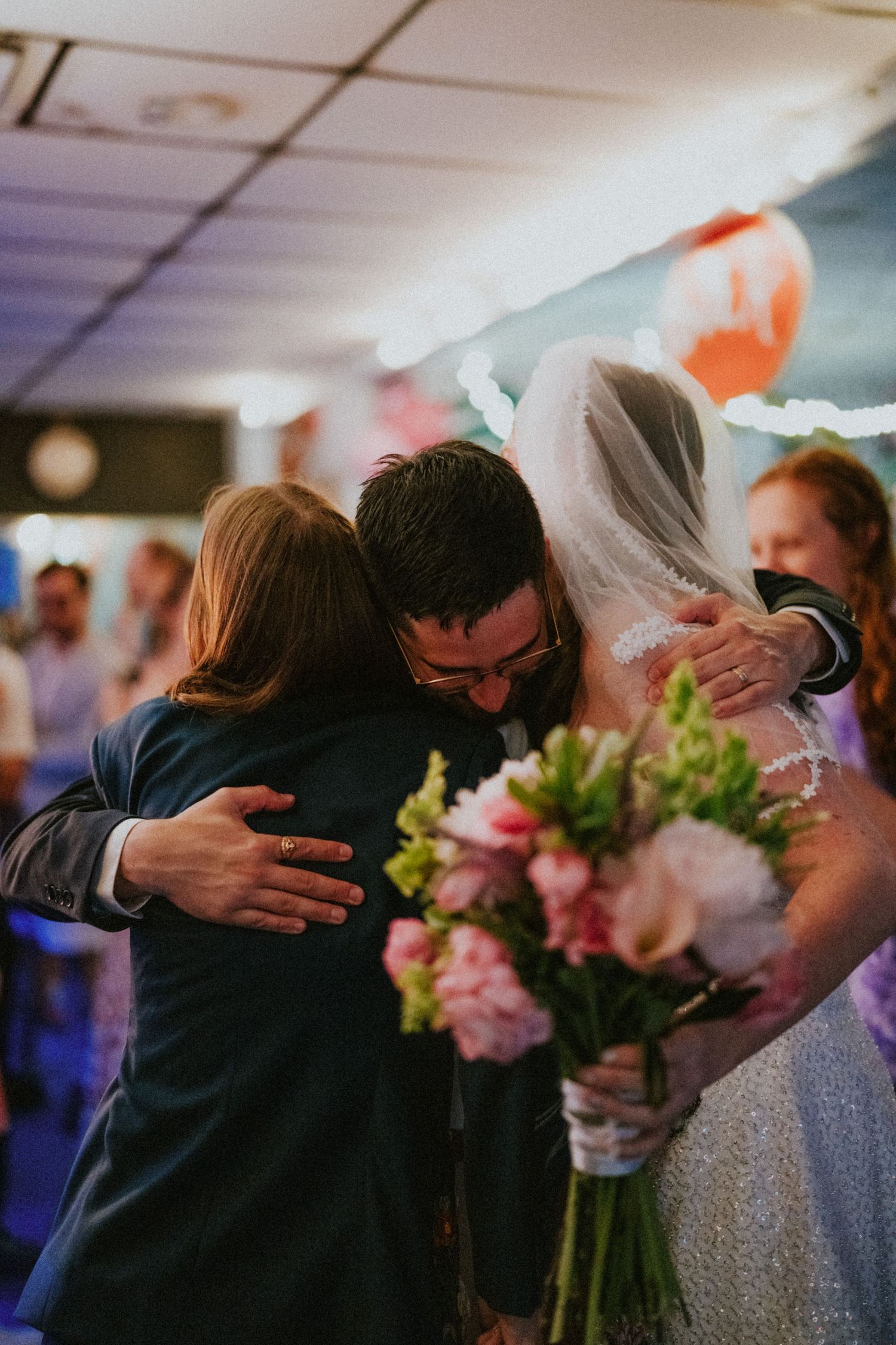 Bride and groom share a group hug with a guest at their wedding reception while bride holds pink bouquet.