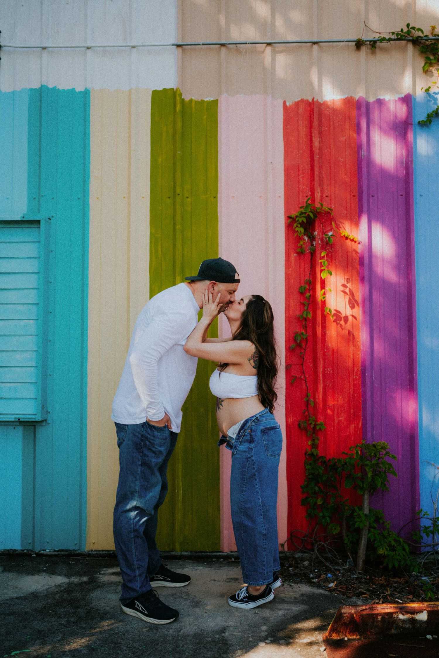A couple embraces against a colorful rainbow striped wall with turquoise shutters in an artistic outdoor photo.