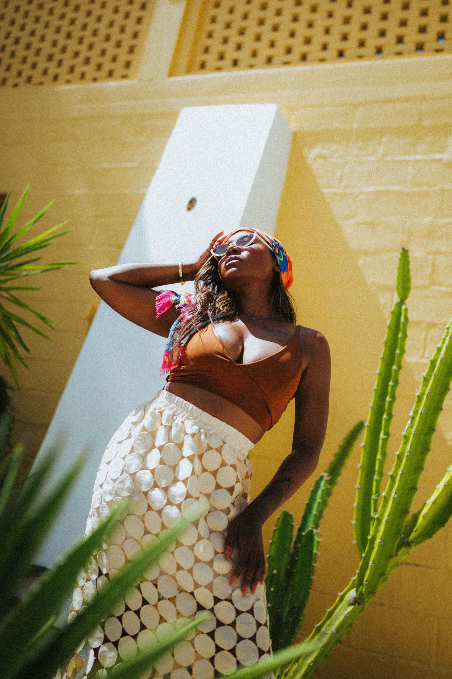 A stylish Black woman poses in a white circle skirt and brown crop top with colorful headscarf against a yellow wall.