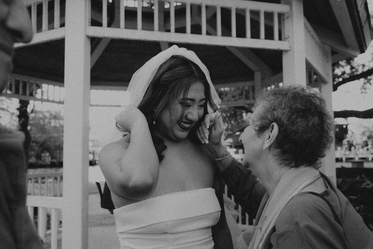Black and white photo of a bride laughing as someone adjusts her veil near a white gazebo outdoors.