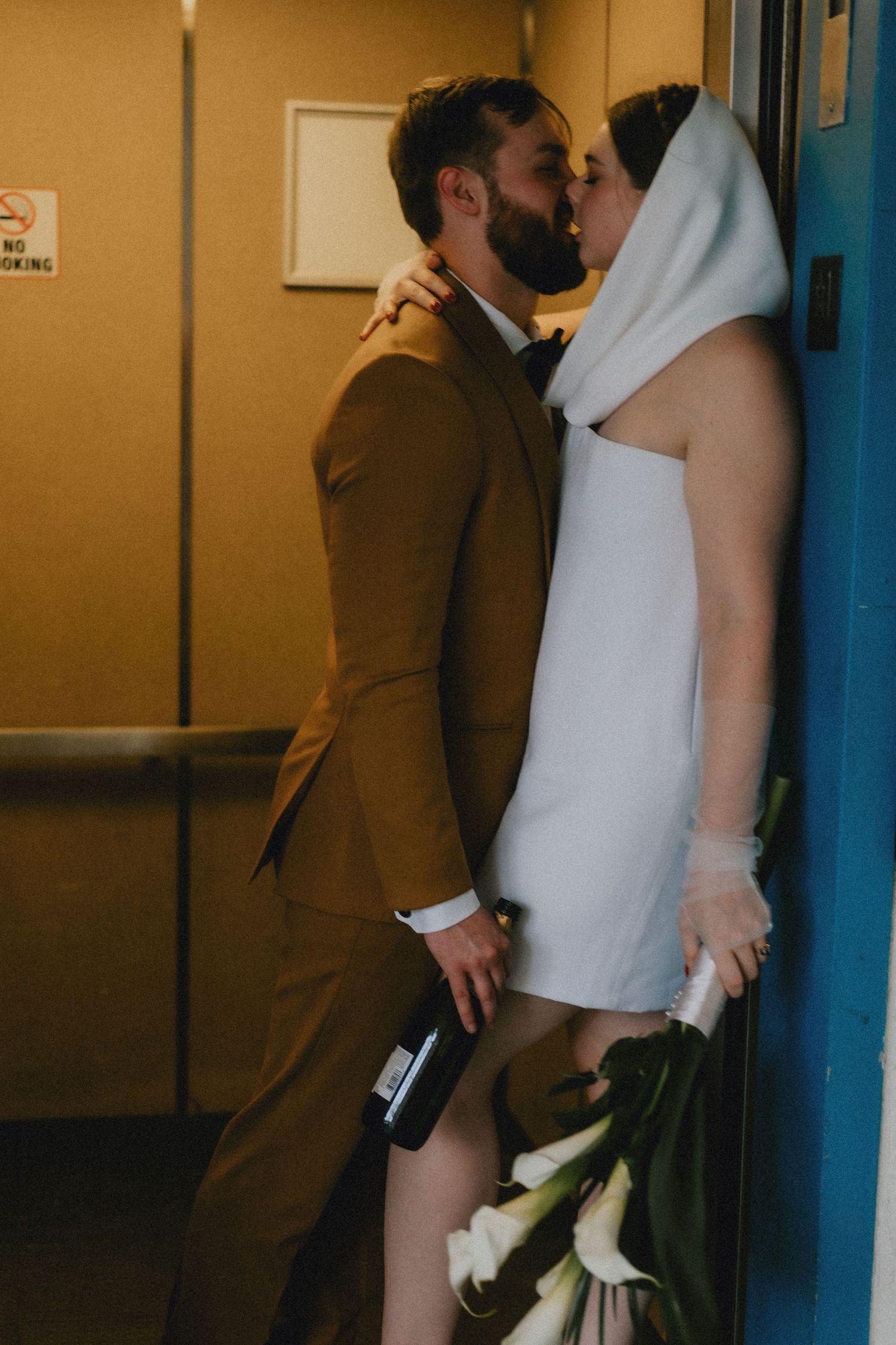 Bride and groom share a romantic kiss in a hallway, she wears a white mini dress with veil, he wears a tan suit.