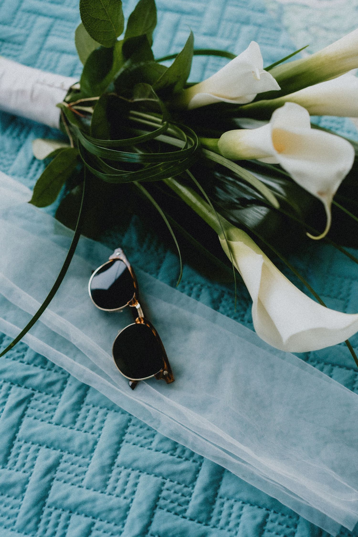 White calla lily bouquet with sunglasses and veil on teal quilted fabric.