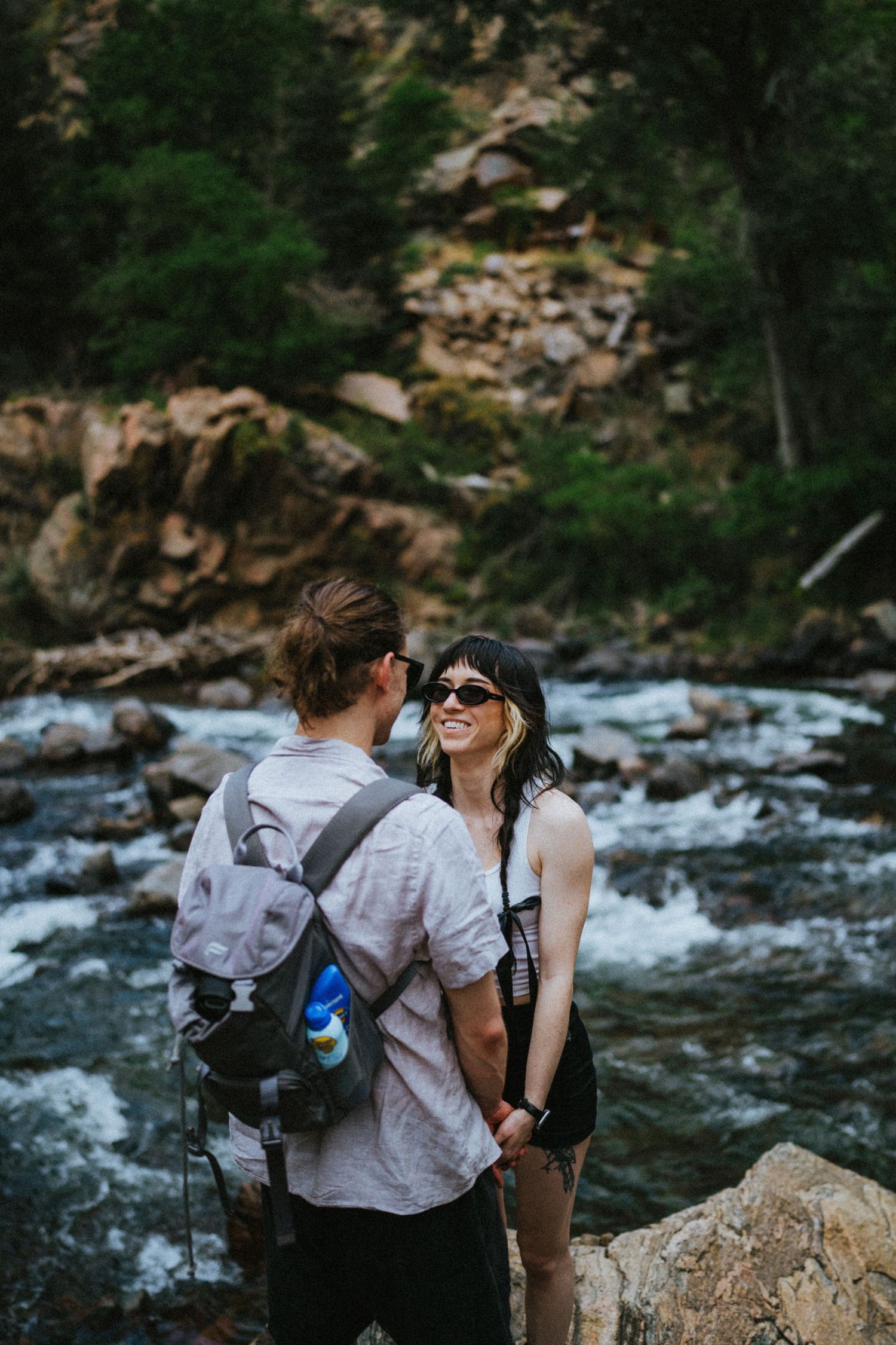 Couple holding hands and smiling by a rushing mountain river surrounded by rocks and forest.