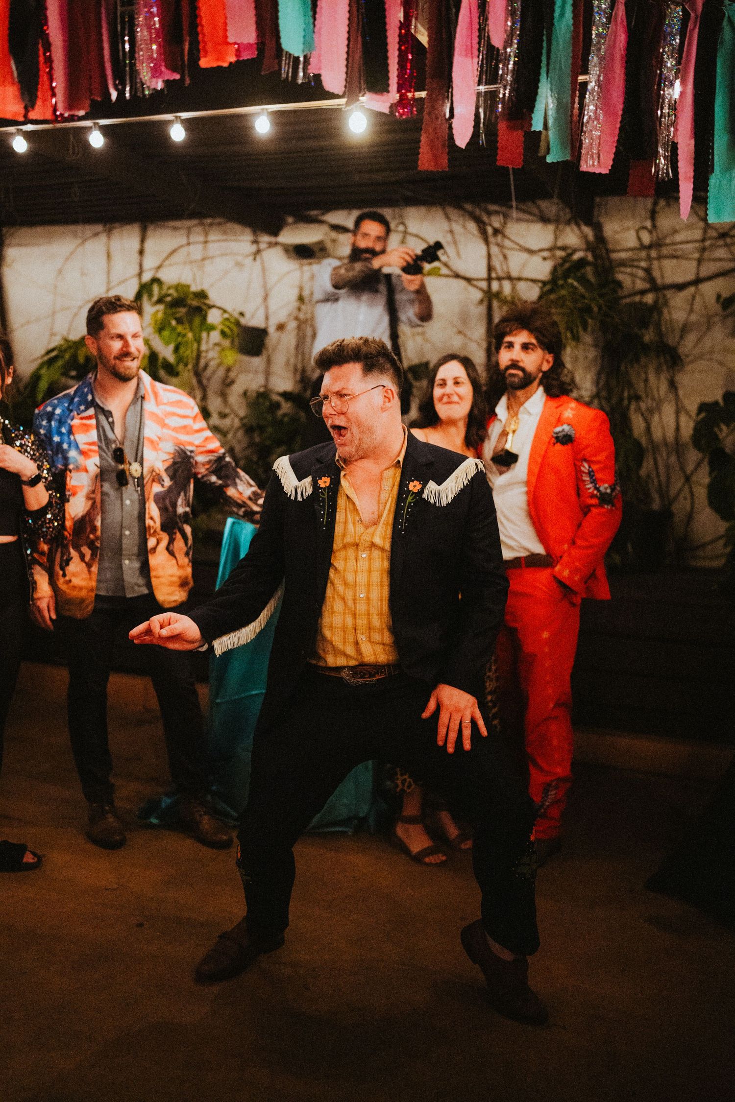 Man in western jacket dancing energetically at a colorful party with guests in vibrant outfits behind him.