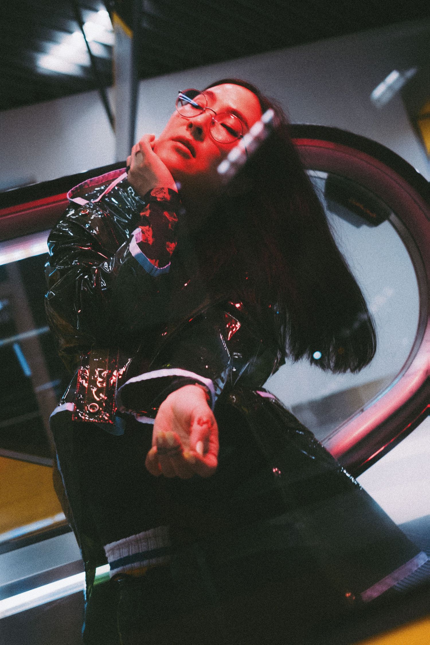 Person in shiny black jacket poses dramatically under red neon lights in a modern urban setting at night.