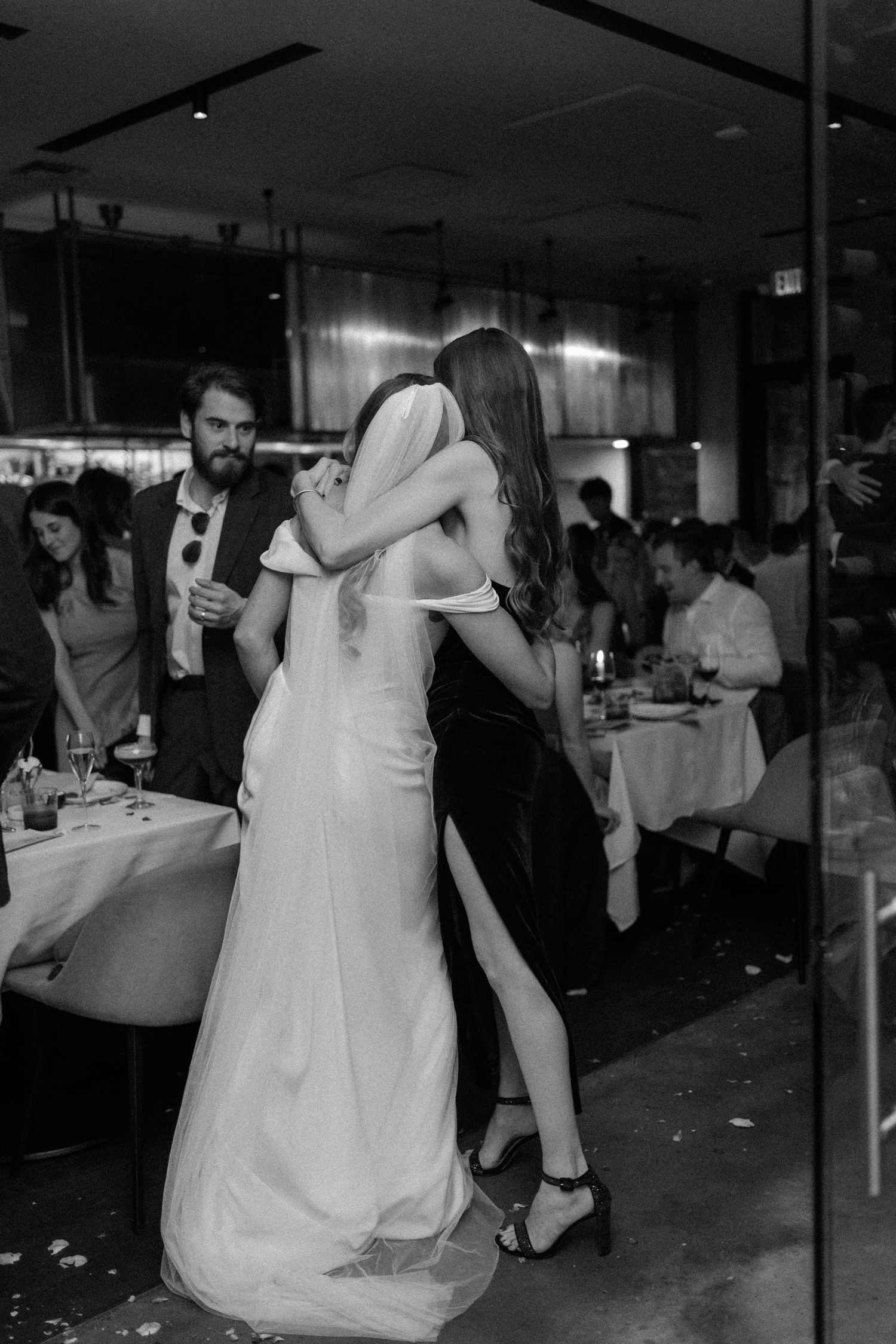 Black and white photo of bride and bridesmaid embracing at wedding reception with guests dining in background.