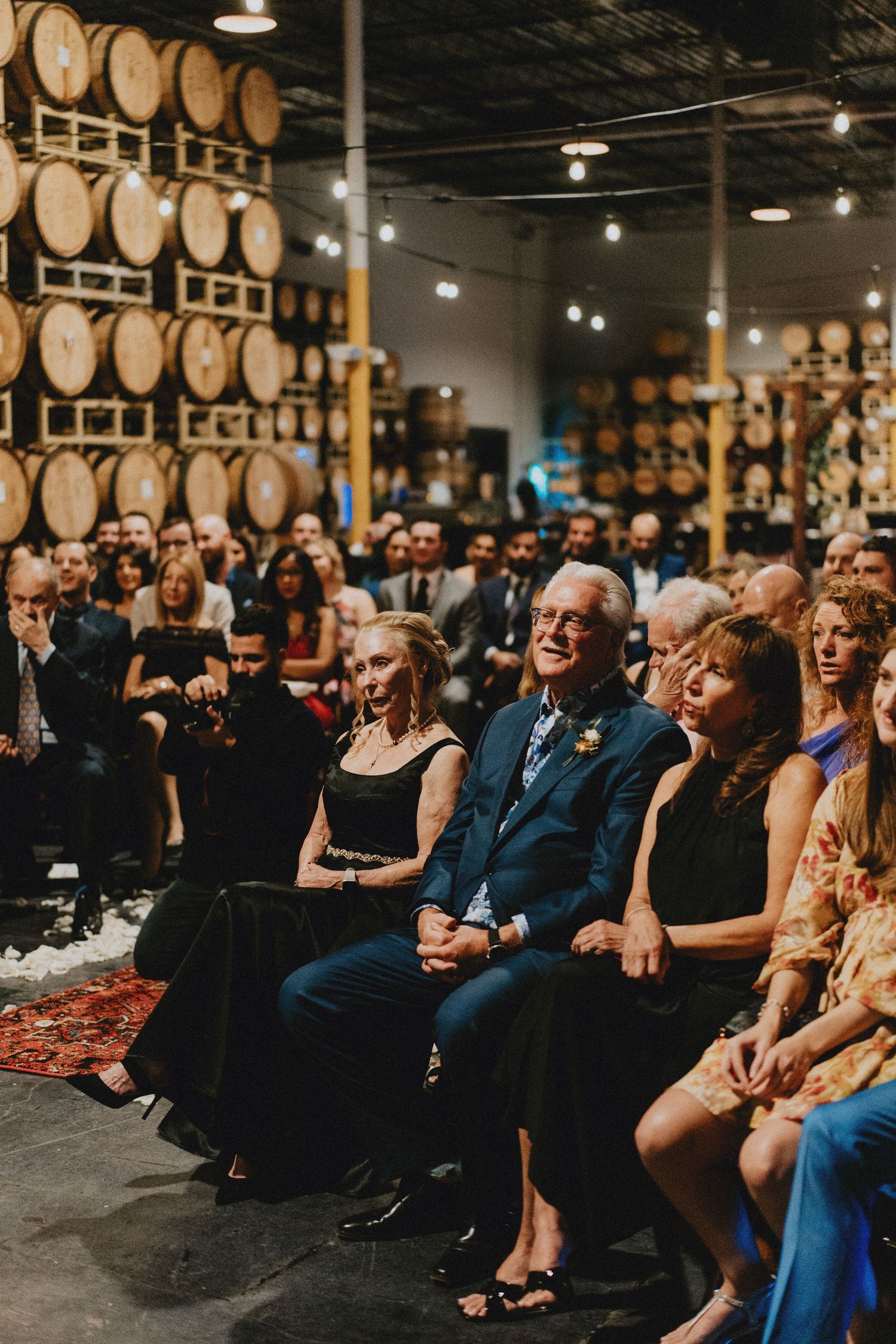 Guests seated at a wedding ceremony inside a rustic brewery with wooden barrels stacked in the background.