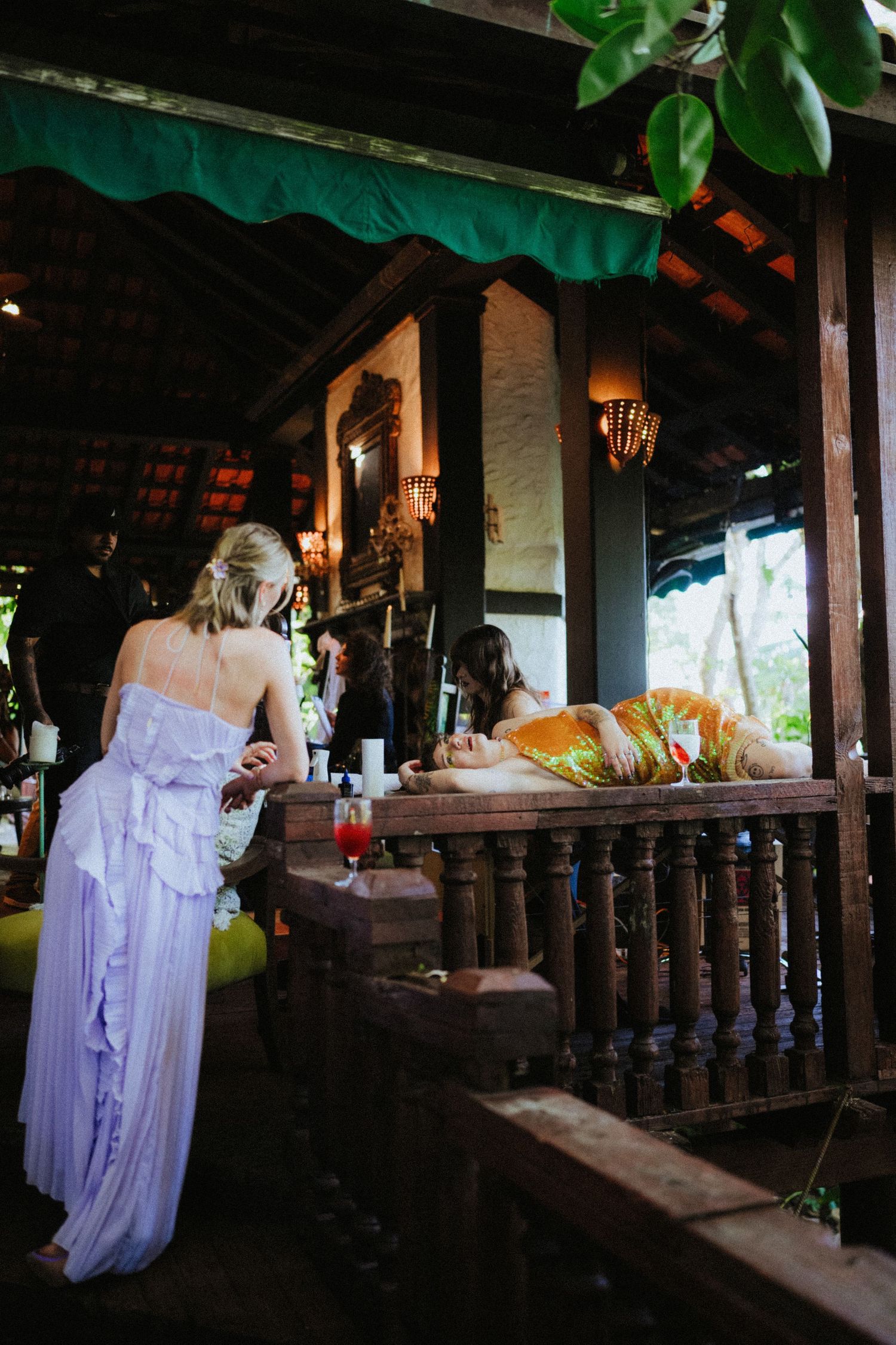 Women in elegant dresses socializing at a rustic tropical bar with wooden railings and warm ambient lighting.