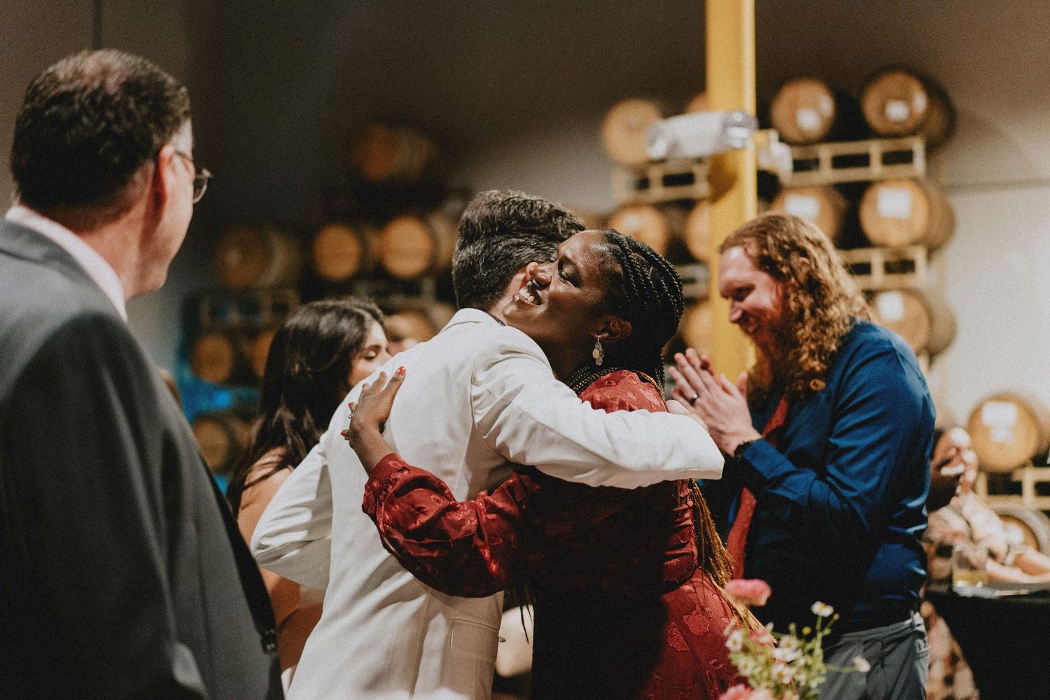 Two people share a warm embrace at a winery event, surrounded by smiling guests and wine barrels in the background.