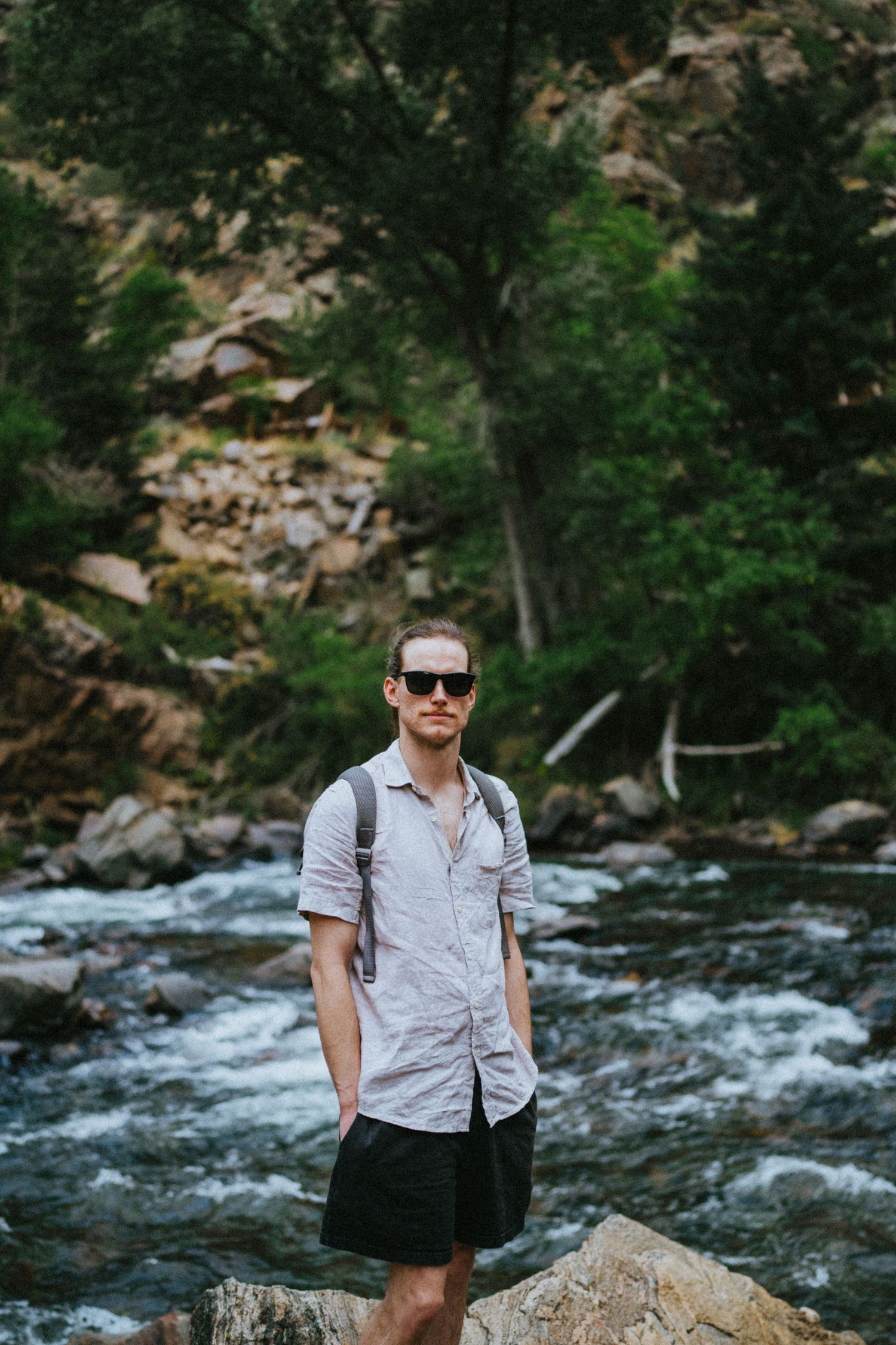 Man in sunglasses and casual shirt stands on rocks by a rushing mountain river surrounded by lush green trees.