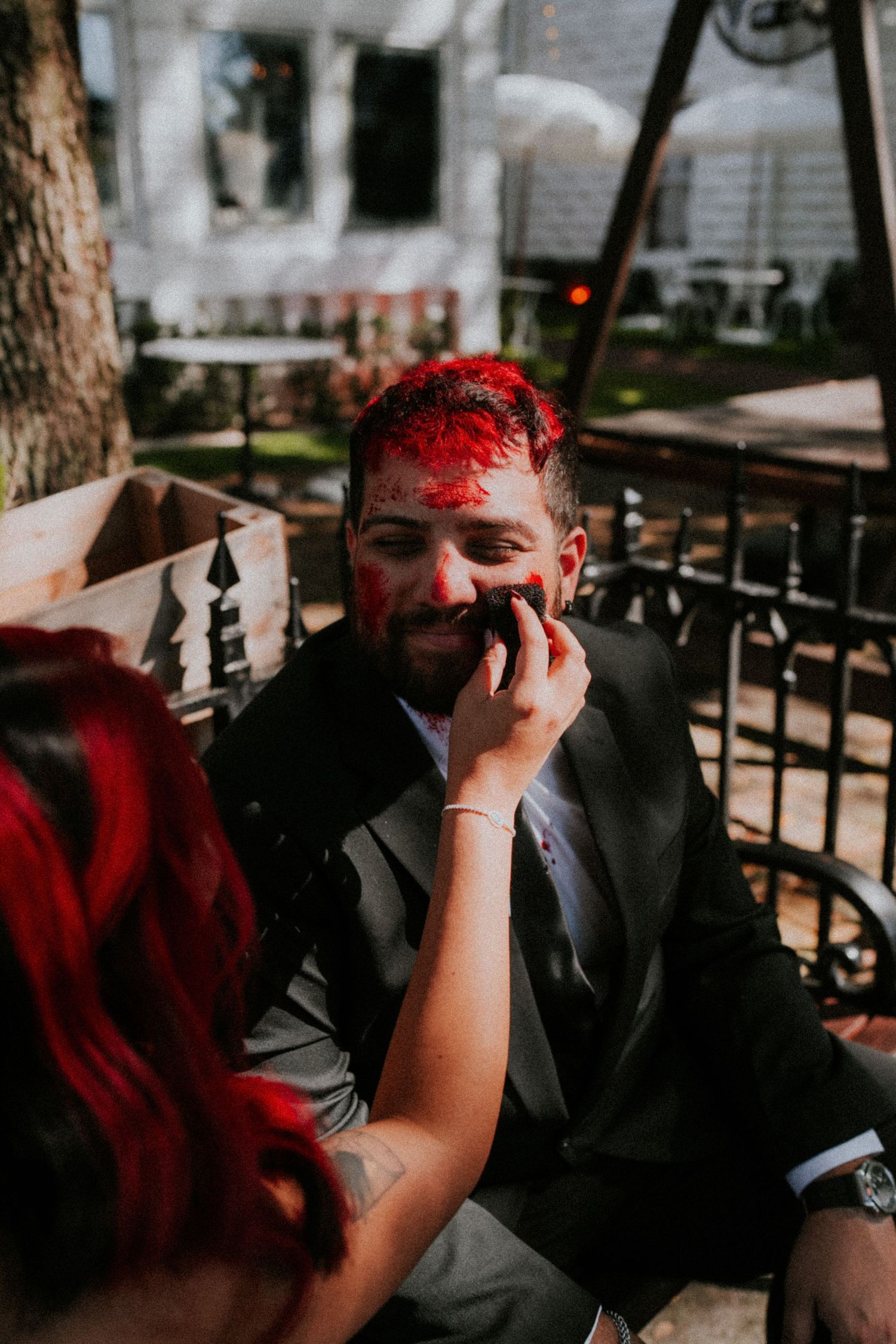 Groom in black suit with red Halloween zombie makeup being applied by red-haired bride outdoors.