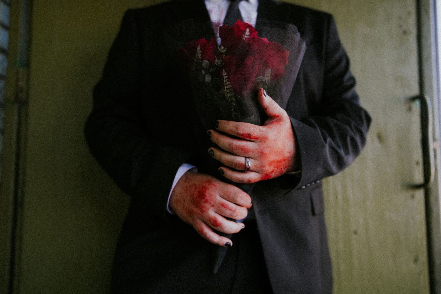 Groom in black suit holding red roses with a wedding ring on his finger.