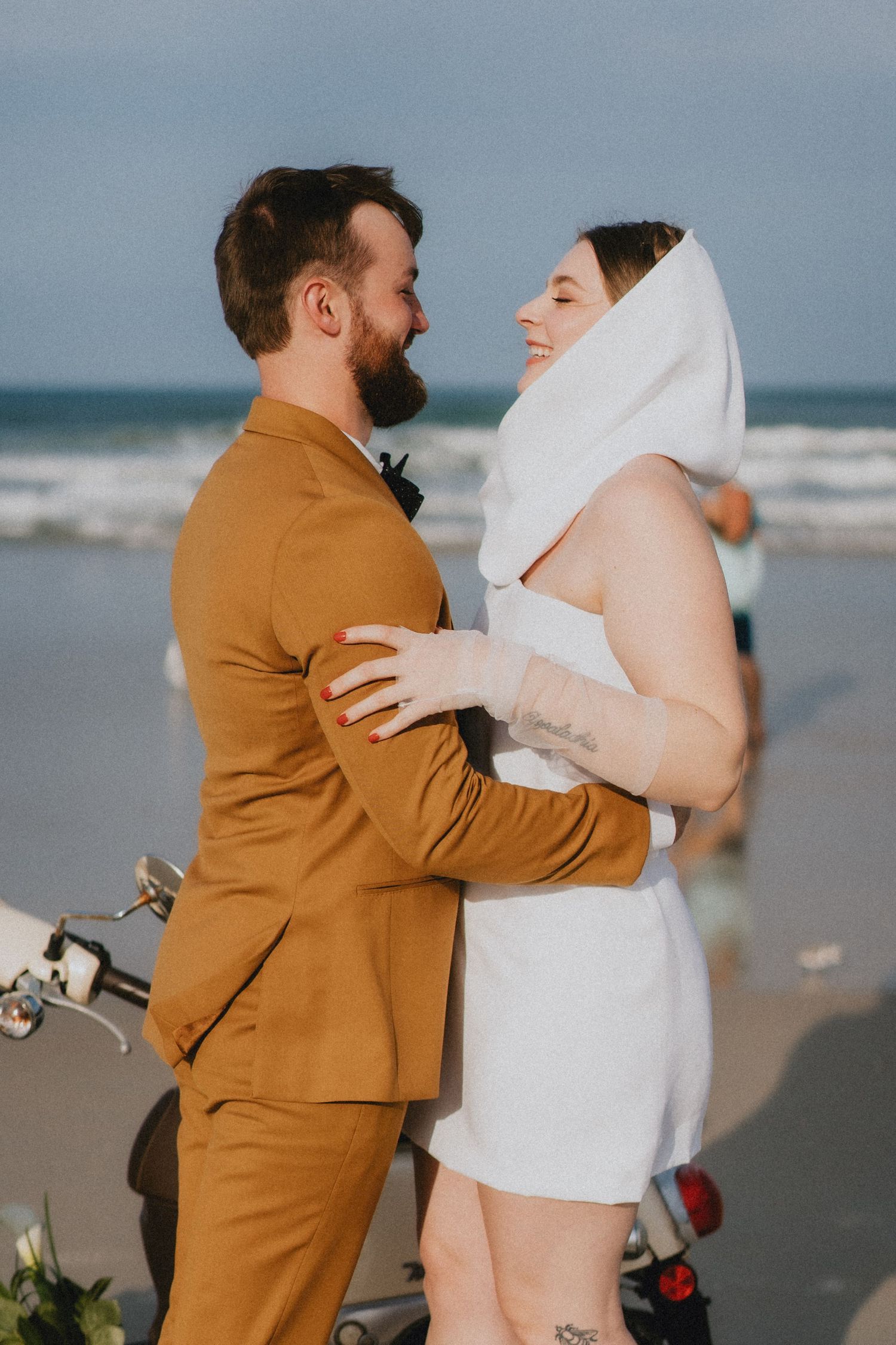 Bride and groom embrace on beach, she wears white mini dress with veil, he wears mustard suit.