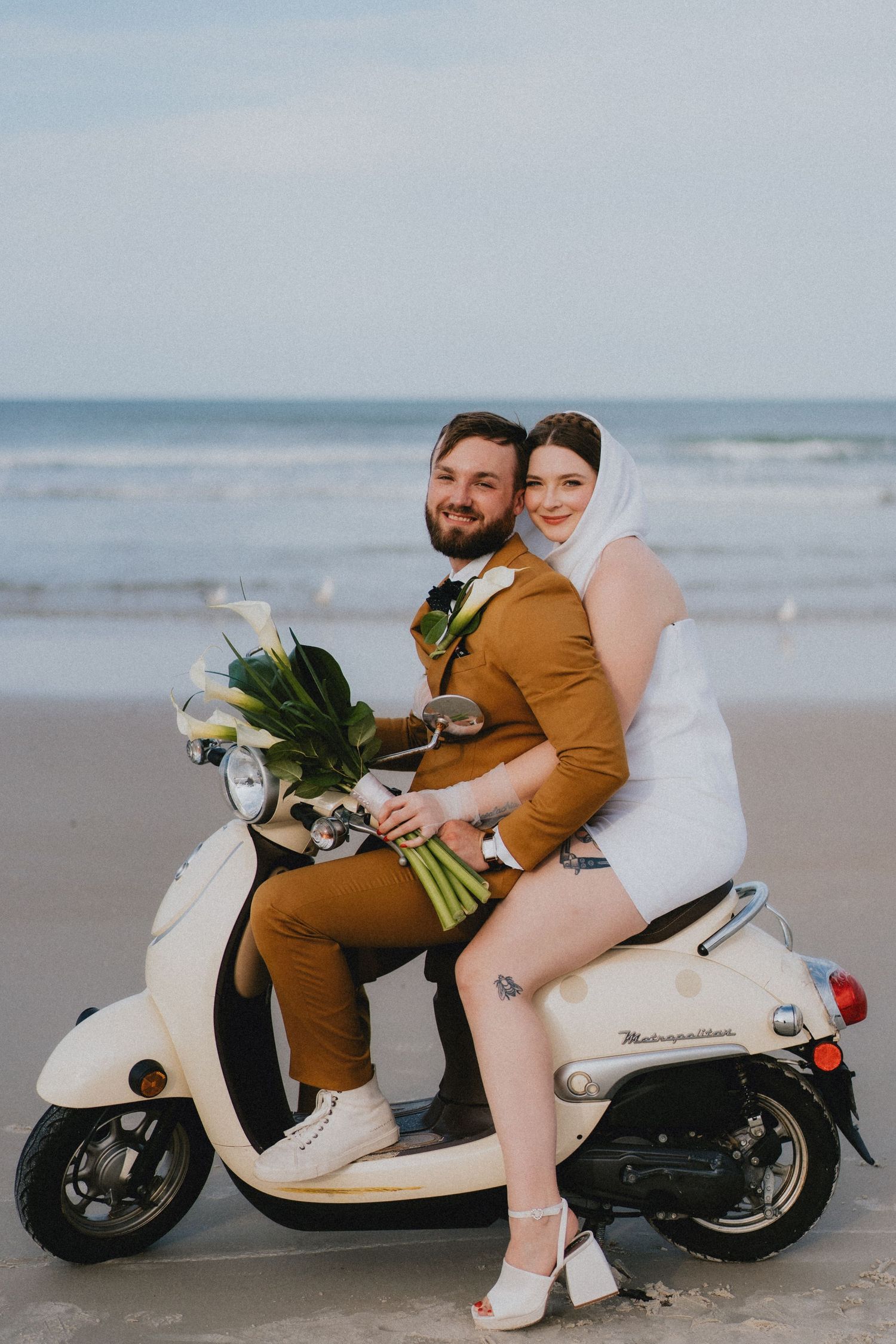 Bride and groom riding a cream Vespa scooter on the beach holding a white calla lily bouquet.