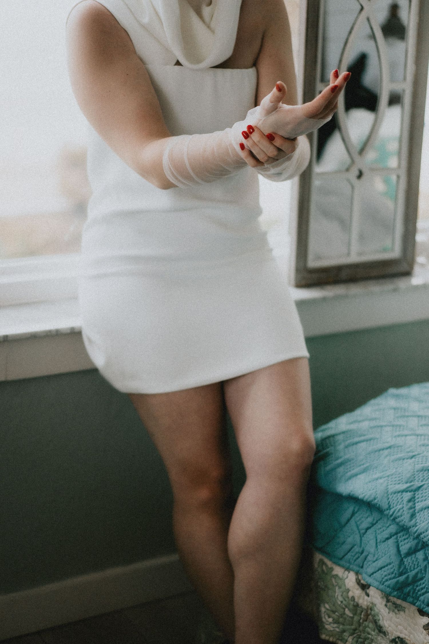 Woman in white mini dress putting on a lace glove with red nails, standing by a window.
