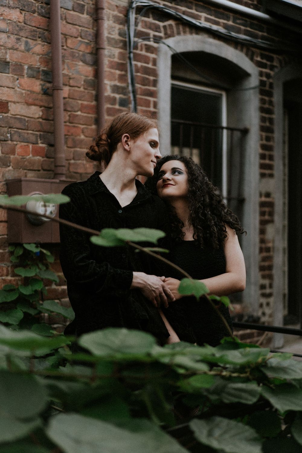 Romantic couple portrait through foliage against brick building.