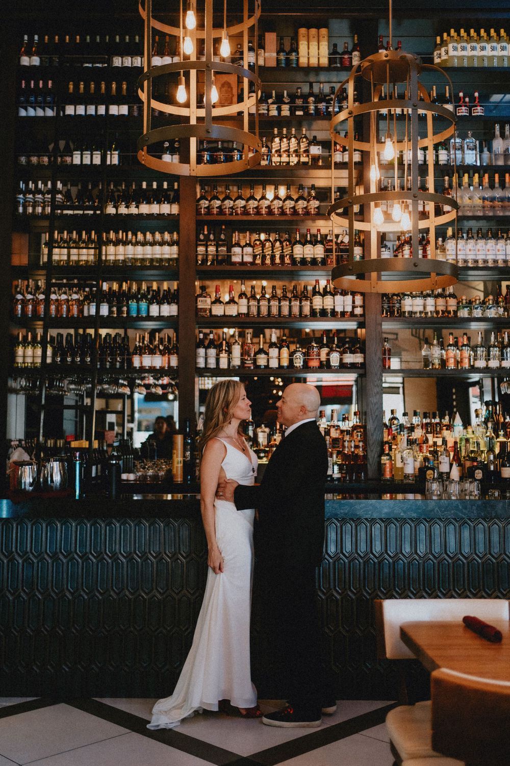 Bride and groom standing at an elegant bar with illuminated shelves of bottles.