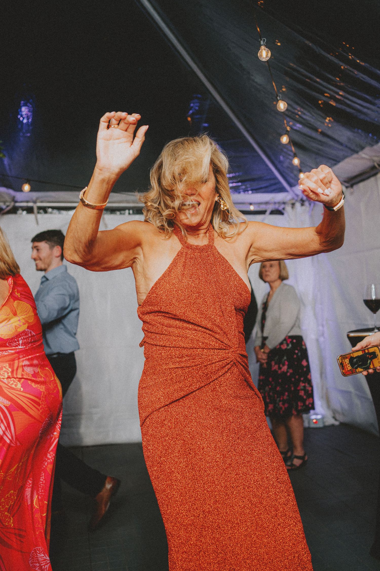 Woman in rust-colored halter dress dances energetically at an outdoor tented wedding reception.