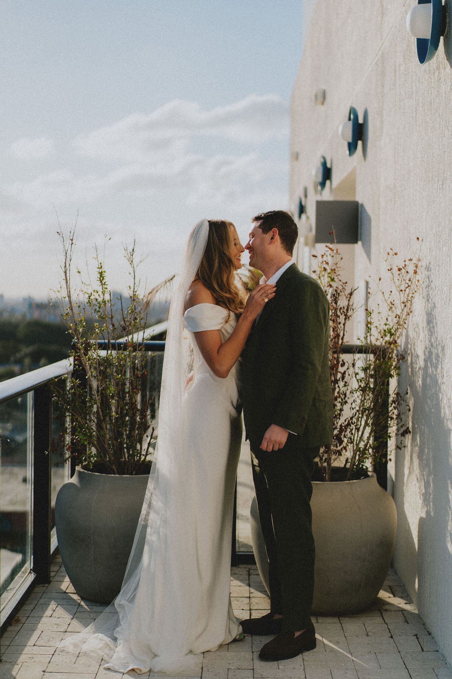 Bride and groom share a romantic kiss on a rooftop balcony, bride wearing a white off-shoulder gown and veil.