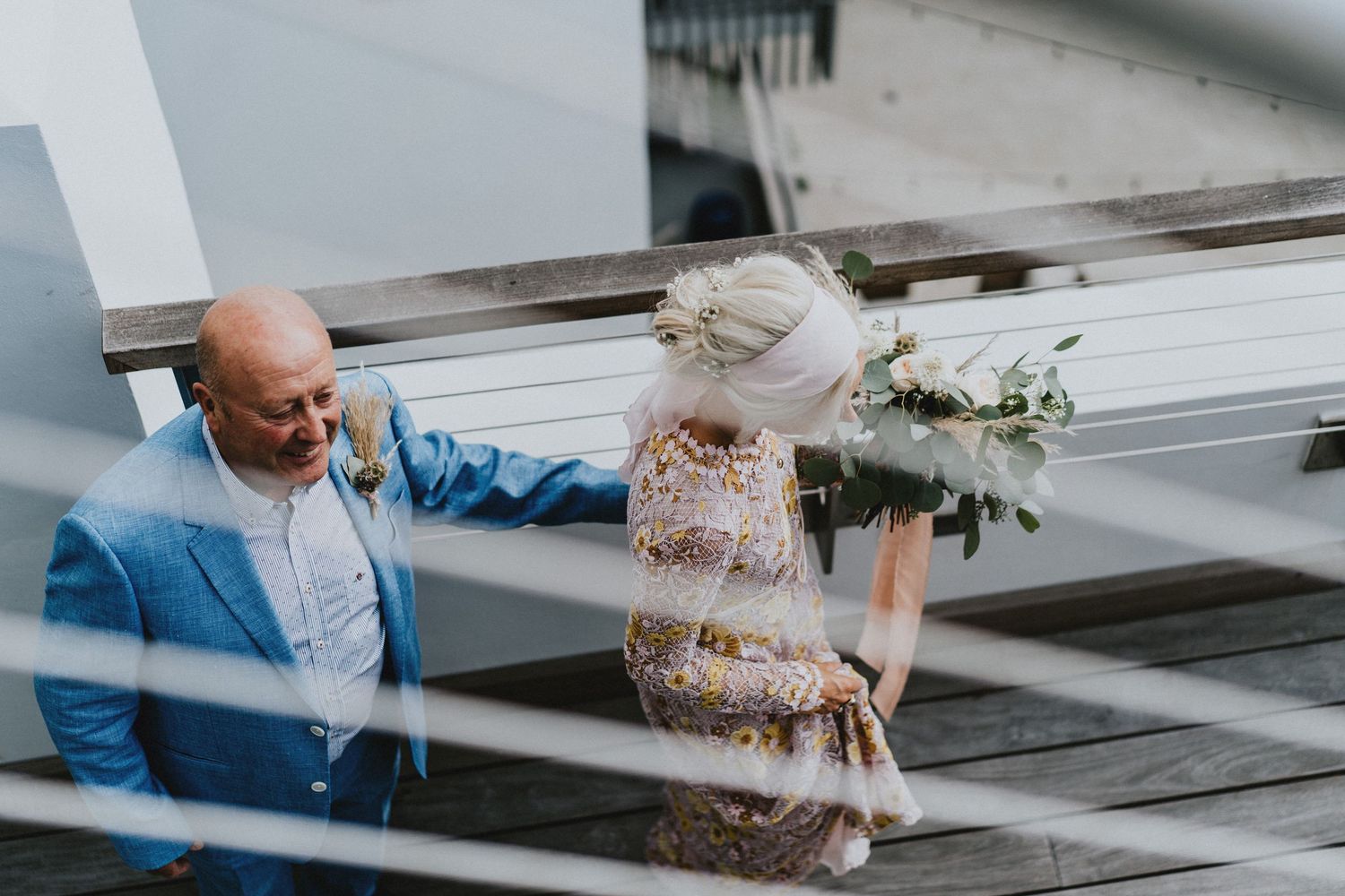Bald groom in blue suit holds bride's hand on wooden deck, bride holding greenery bouquet, aerial view.