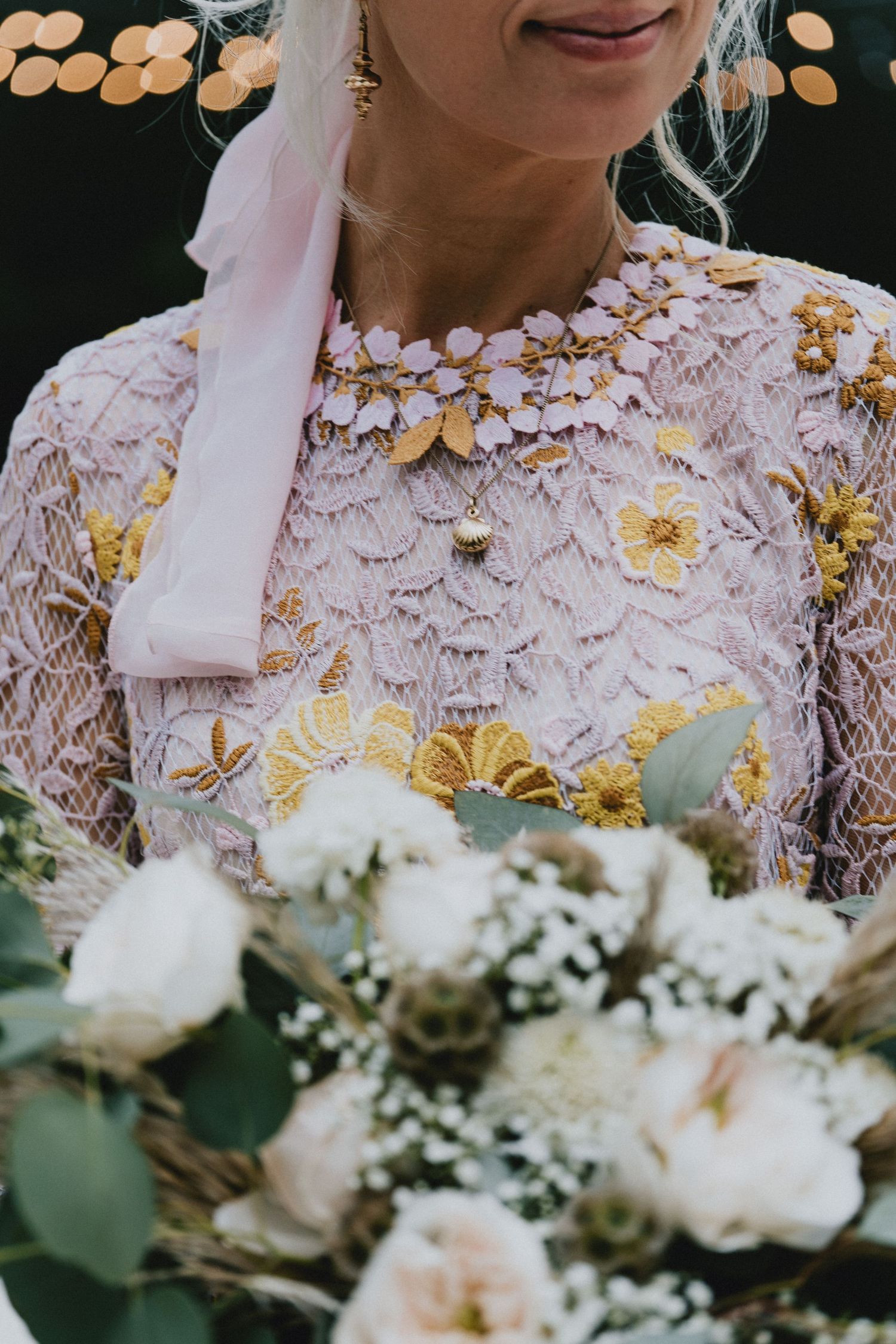 Bride in pink lace dress with gold floral embroidery holds white rose bouquet, wearing matching floral necklace.