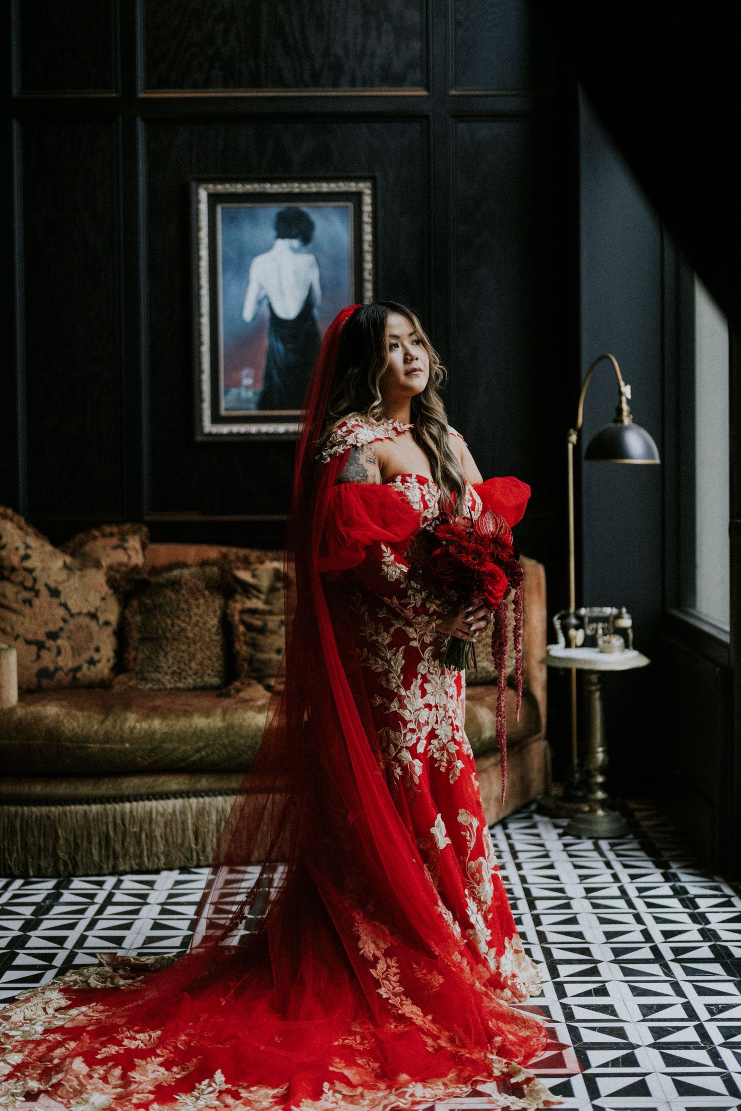 Stunning bride in red floral lace gown with long red veil holding bouquet in dark elegant room.