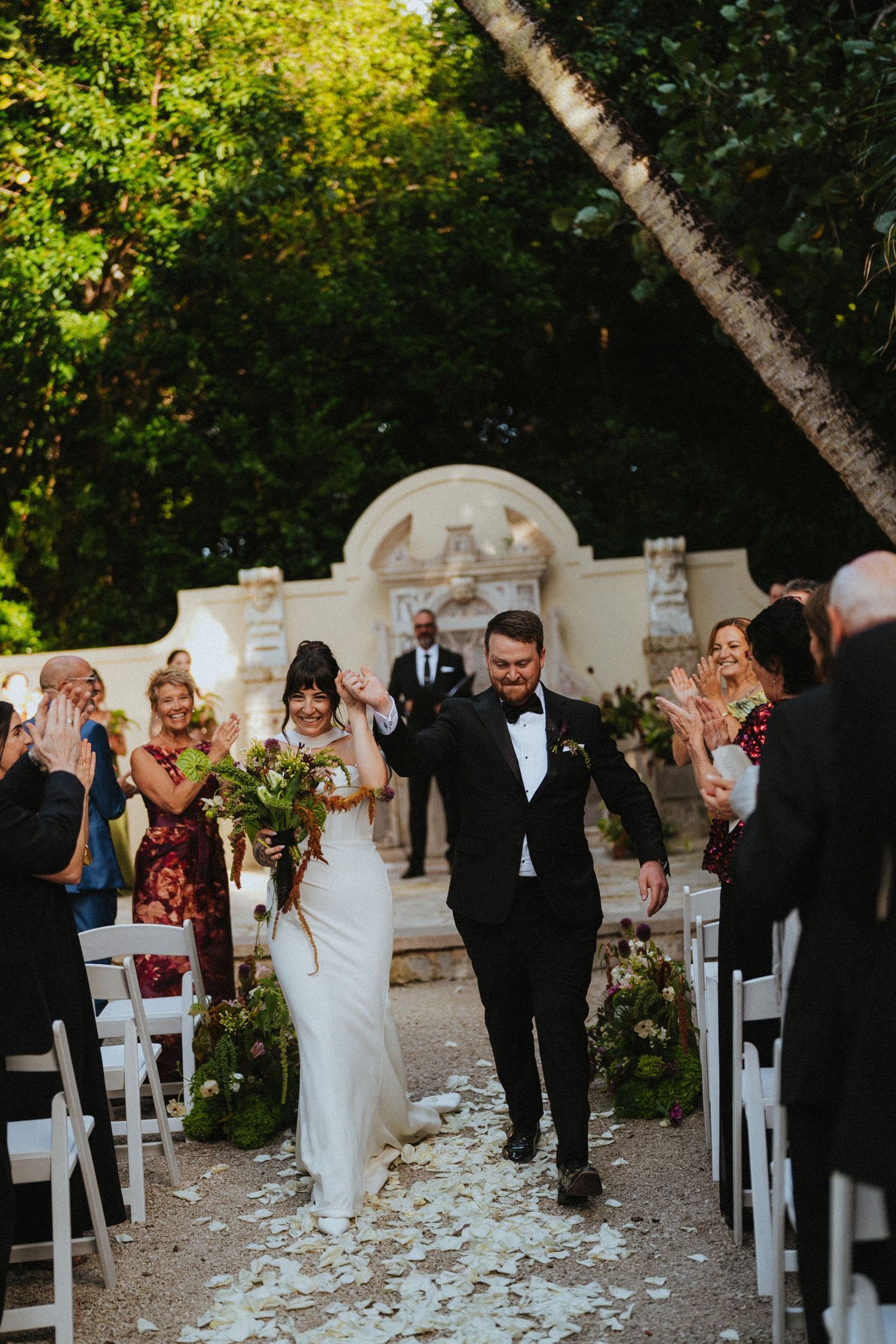Newlyweds walk triumphantly down the aisle as guests celebrate with flower petals at an outdoor garden wedding.