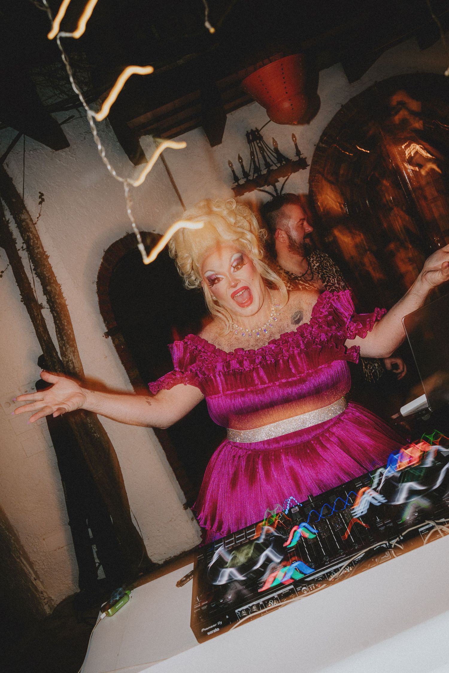 Drag queen in pink ruffle outfit DJing energetically at a lively nightclub event with neon lights.