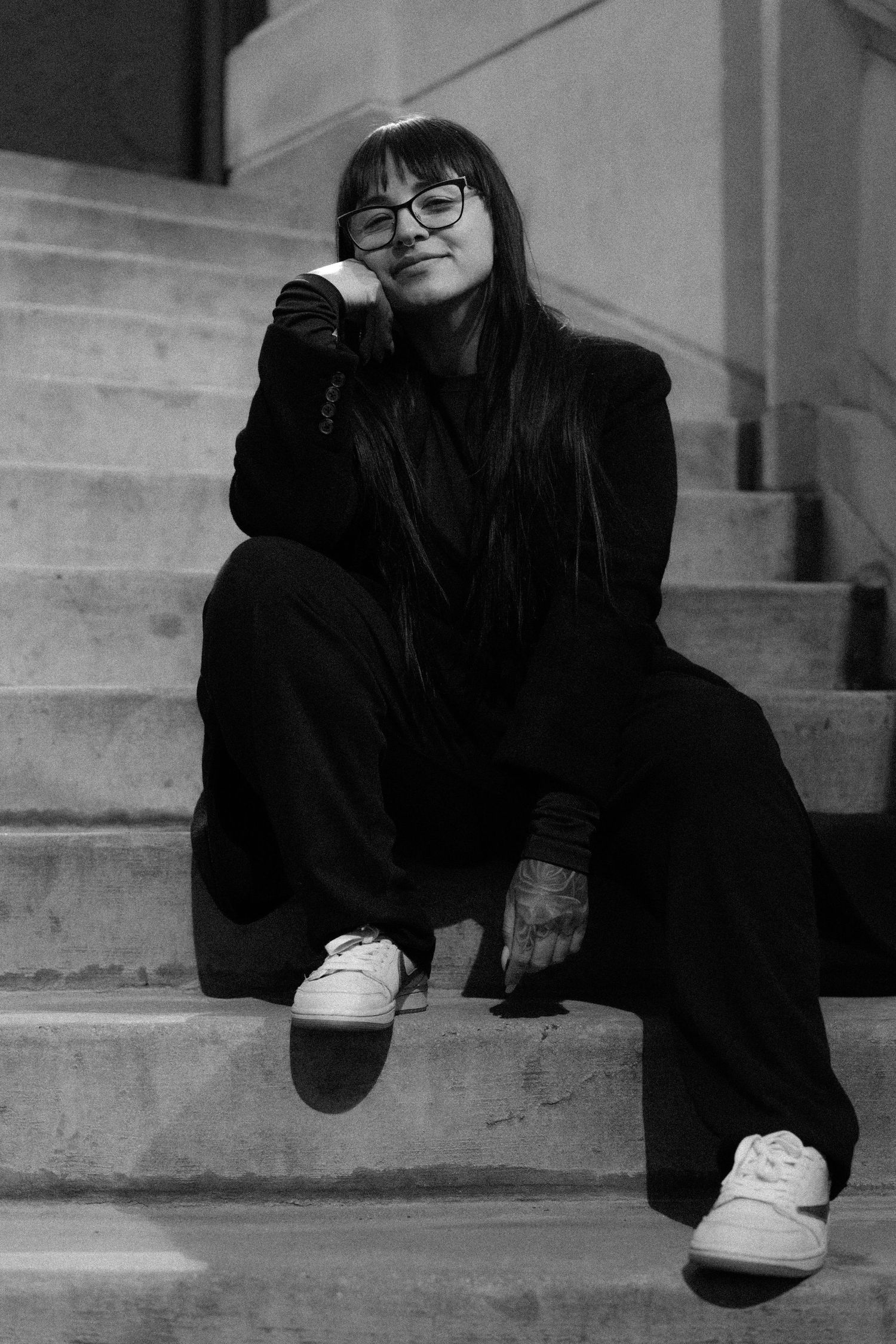 Black and white photo of a young woman in glasses and all-black outfit sitting on concrete steps, smiling softly.