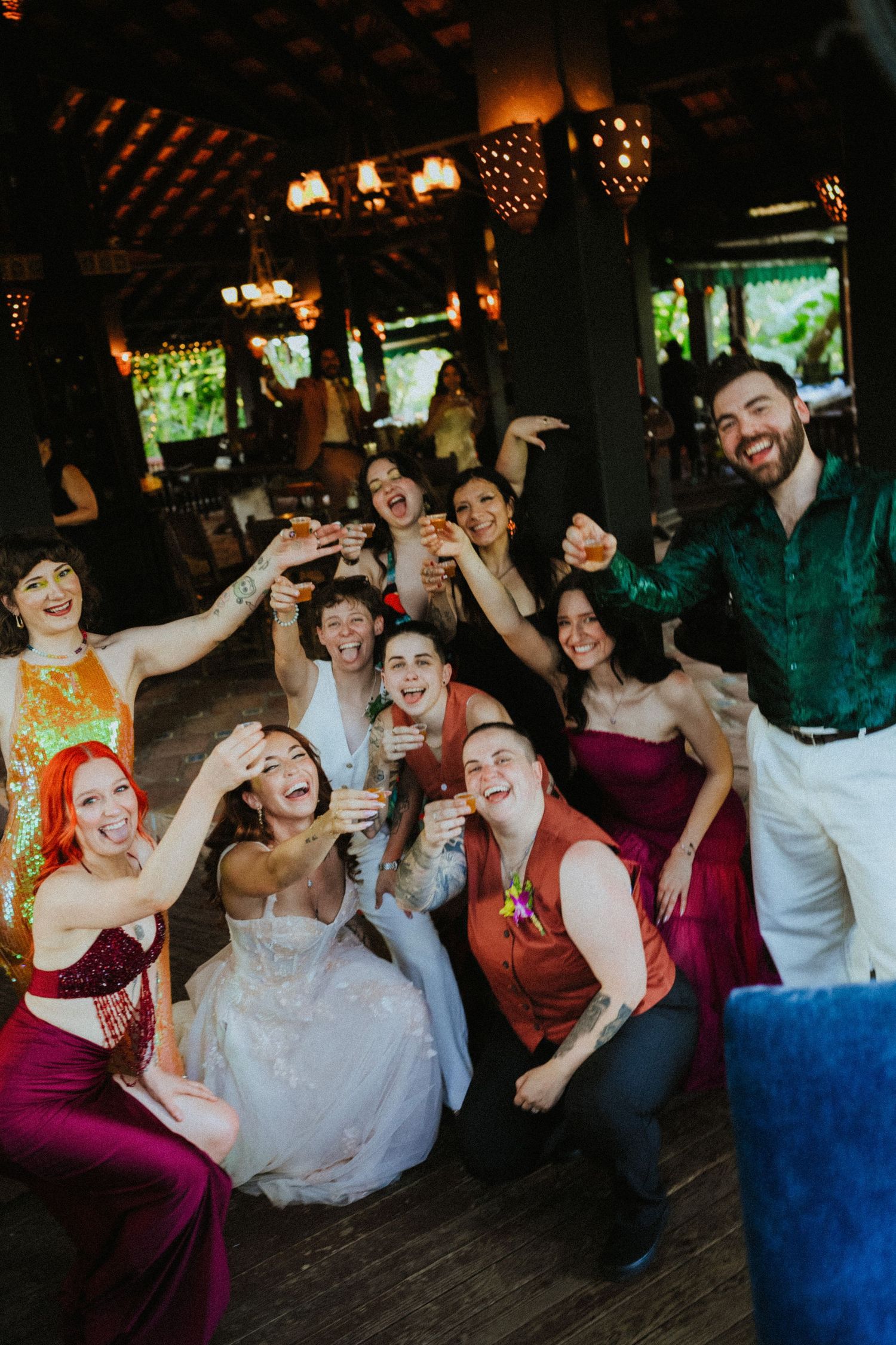 A joyful wedding group raises glasses in a toast, with the bride in white surrounded by colorful-dressed guests at a reception.