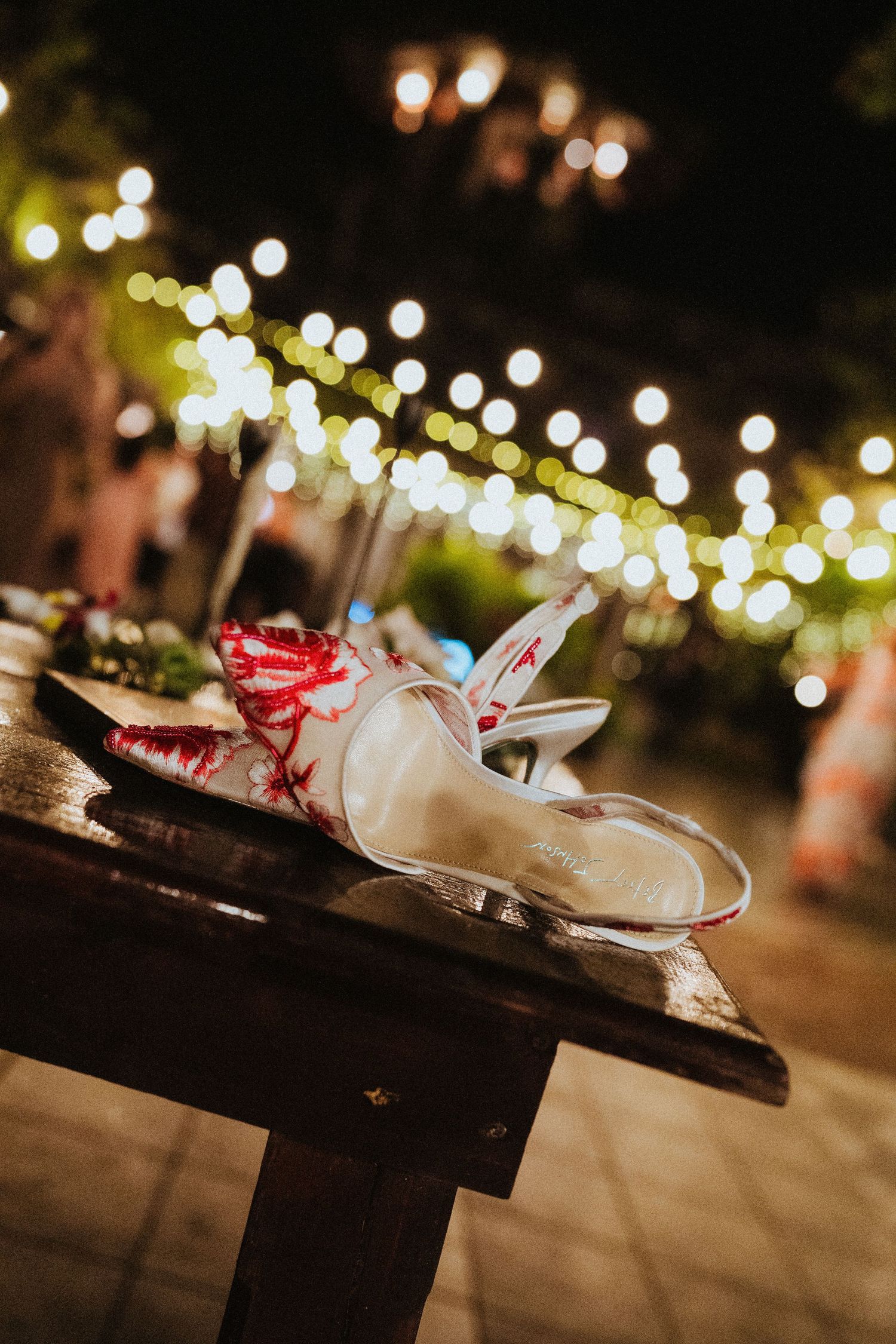 White heeled sandals with red floral fabric resting on a wooden bench at an outdoor evening event with string lights.